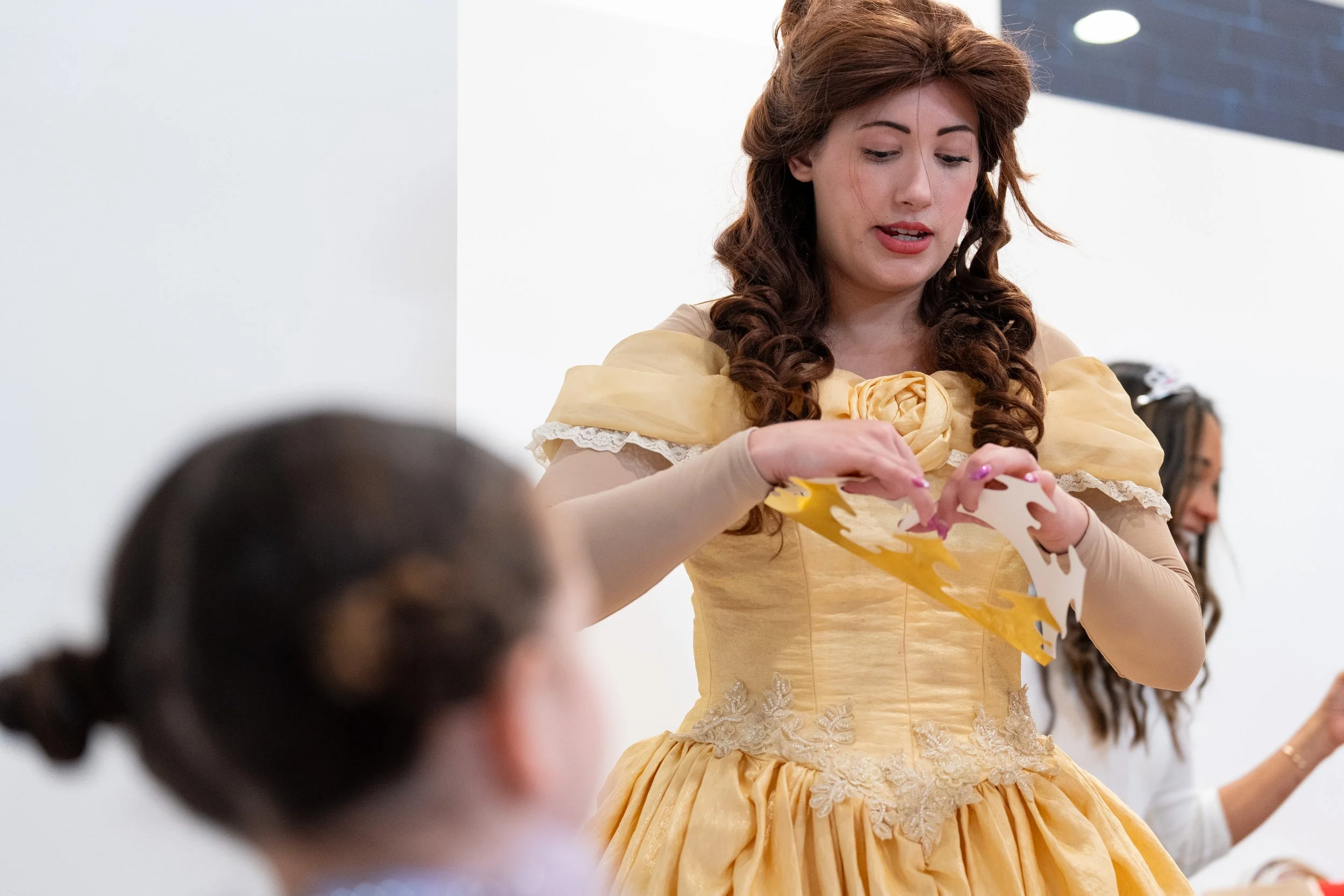 A woman dressed as Belle from Beauty and the Beast, wearing a yellow ball gown, is placing a paper tiara on a girl with dark hair in a bun.
