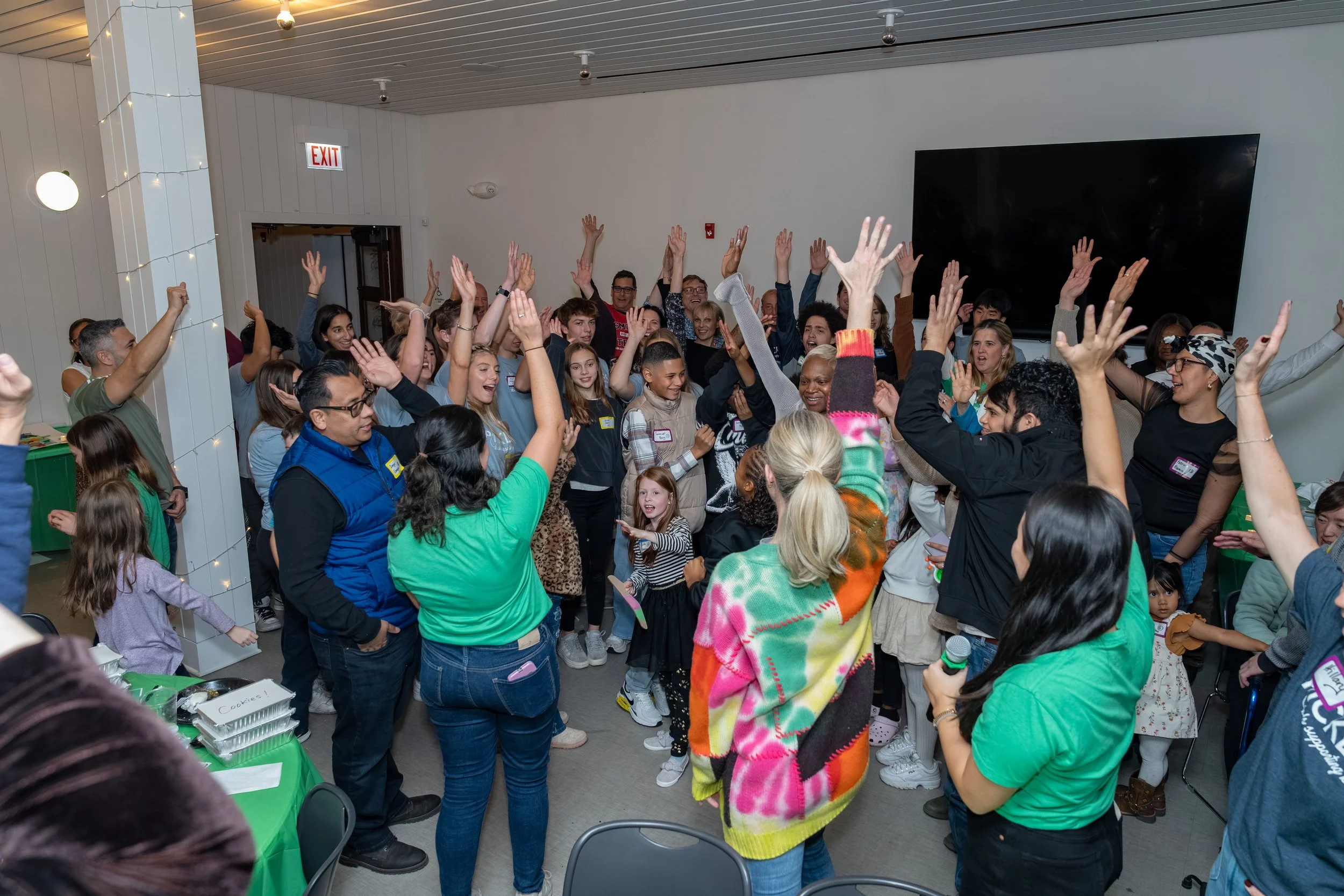 A large group of diverse people, including children and adults, are gathered in a room, raising their hands and dancing together at a celebration or party.