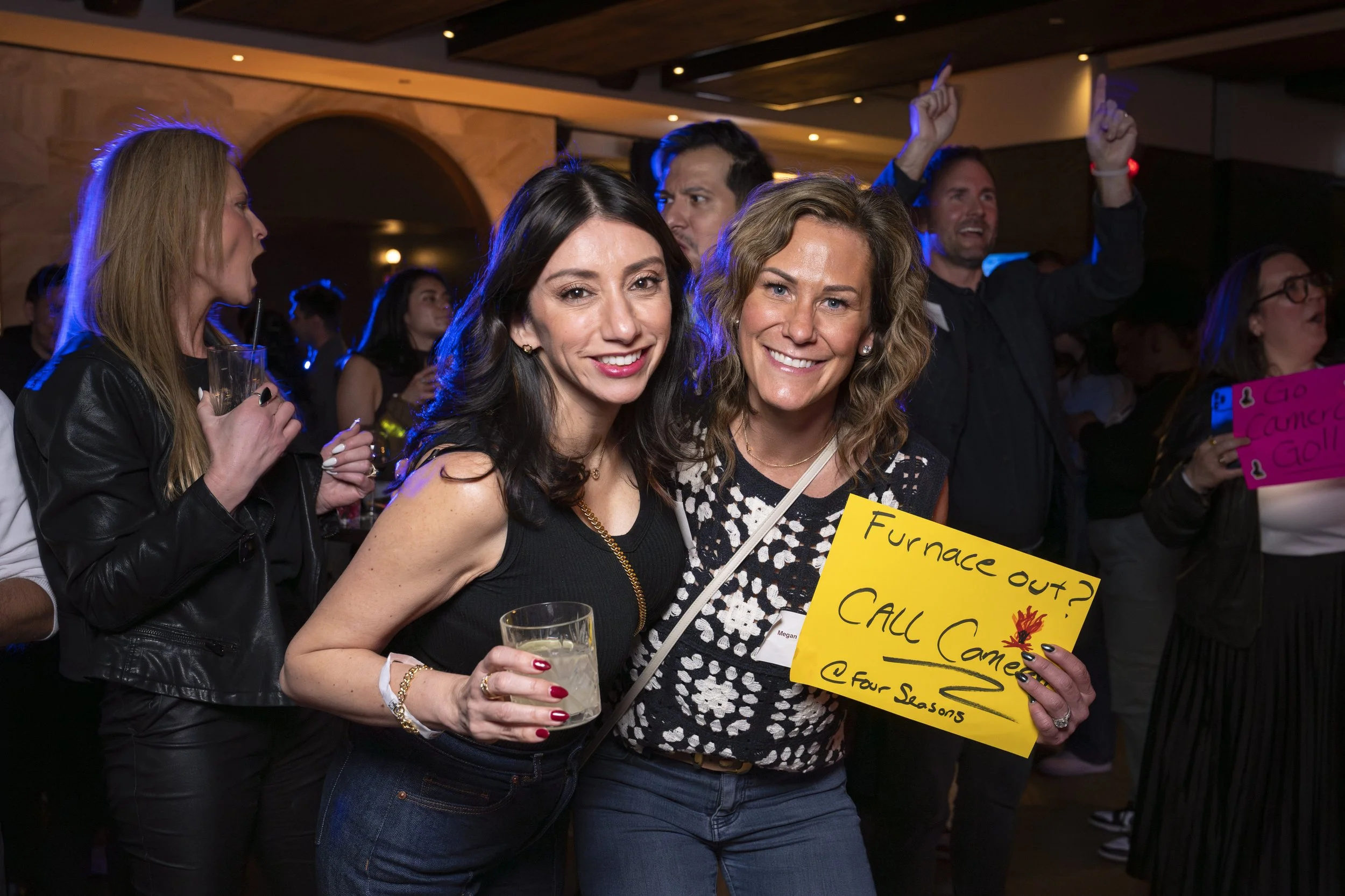 Two women smiling at a party, one holding a drink, the other holding a yellow sign reading 'Furnace out? Call Came' with a flame drawing, surrounded by other party guests.