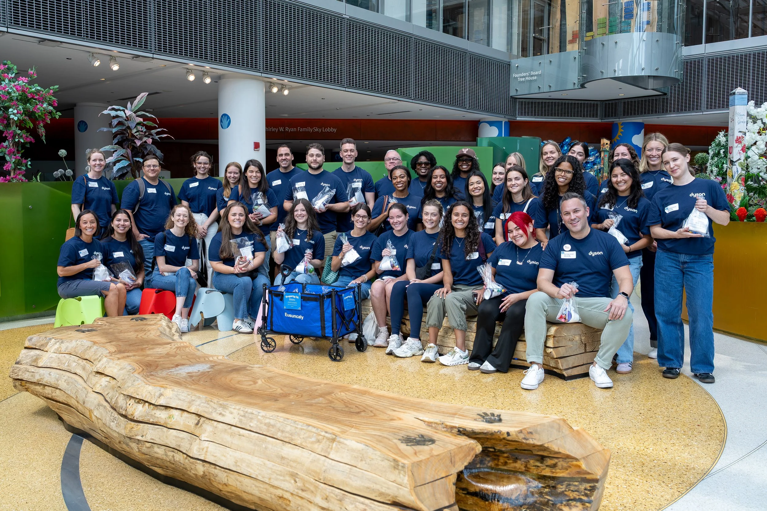 Group of smiling people wearing matching blue shirts gathered in an indoor space, some holding small bags, with a large wooden bench in front and colorful plants around.