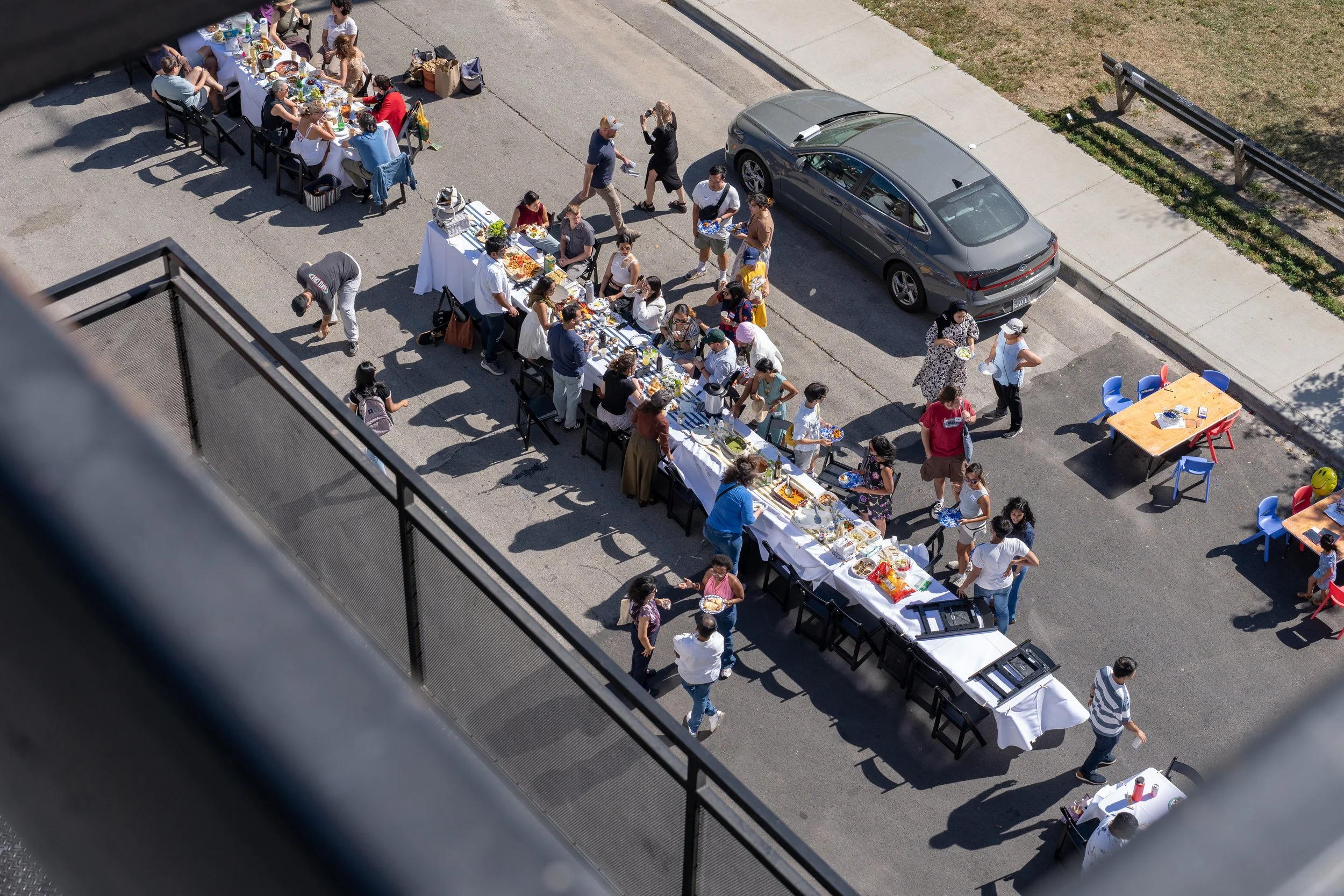 An outdoor gathering with many people standing in line at a long buffet table filled with food. Several other tables with chairs are set up on the street, with some people sitting or walking nearby. A car is parked next to the sidewalk, and the scene