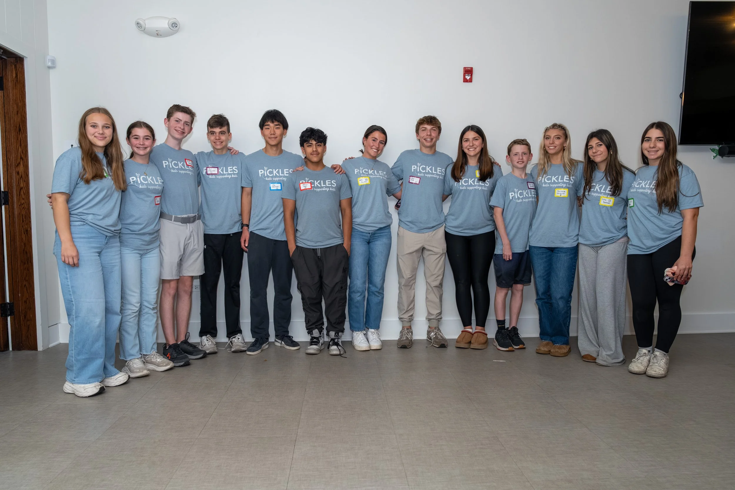 Group of young people posing together indoors, all wearing matching blue T-shirts with the word 'PICKLES' printed on them, smiling at the camera.