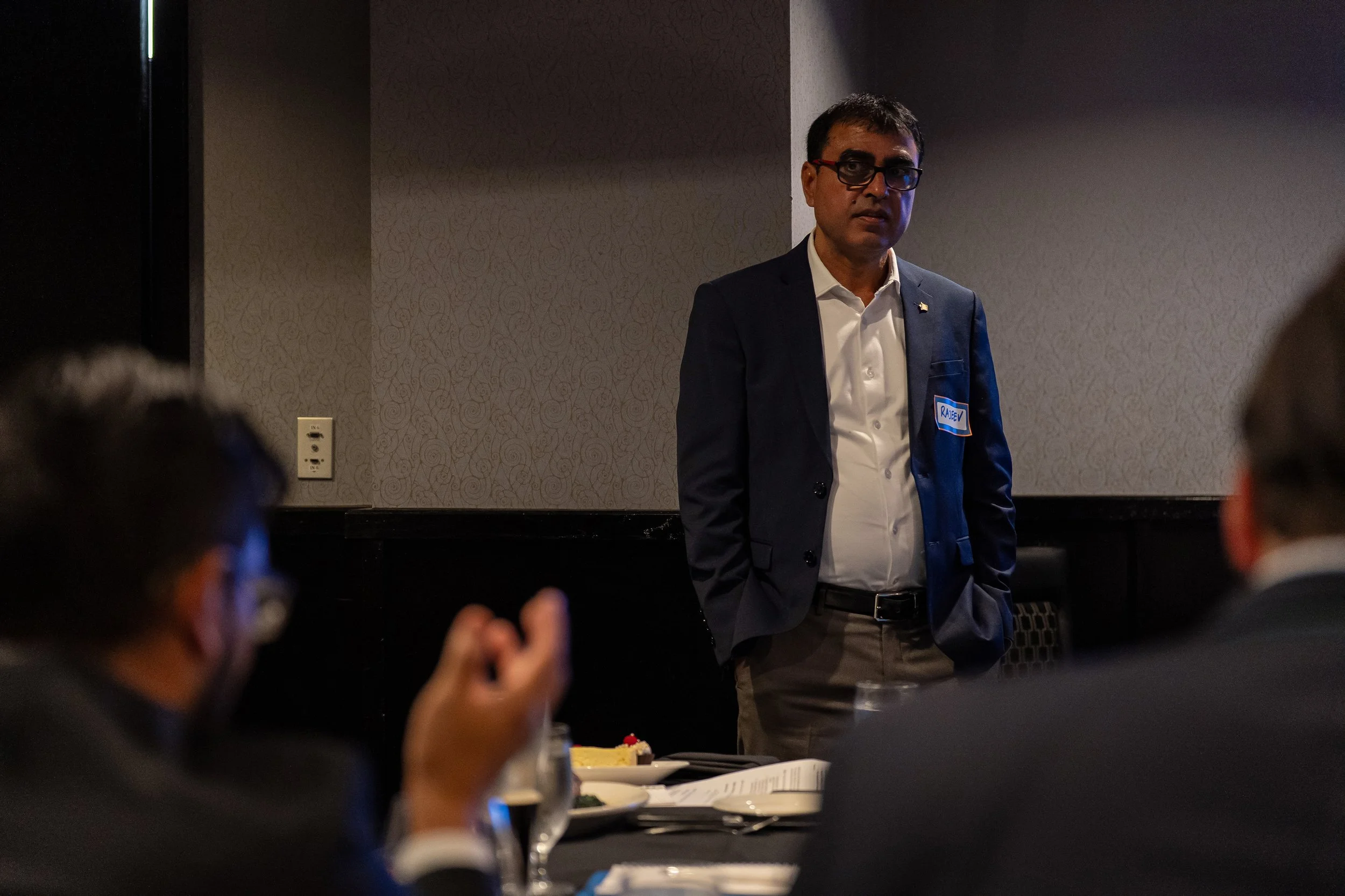 Man wearing glasses and a dark suit standing in front of a gray patterned wall at a conference or meeting, with seated people around a dining table in the foreground.