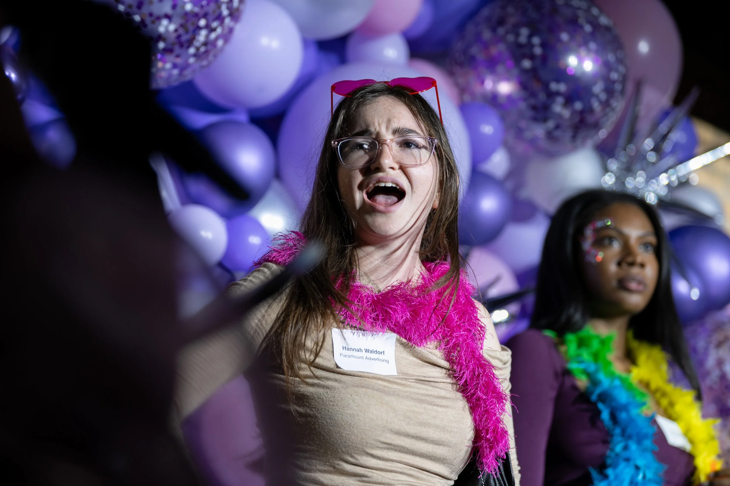 A young woman with long brown hair, glasses, and a pink feather boa expresses surprise in front of purple balloons, with another woman beside her also wearing colorful accessories at a festive event.