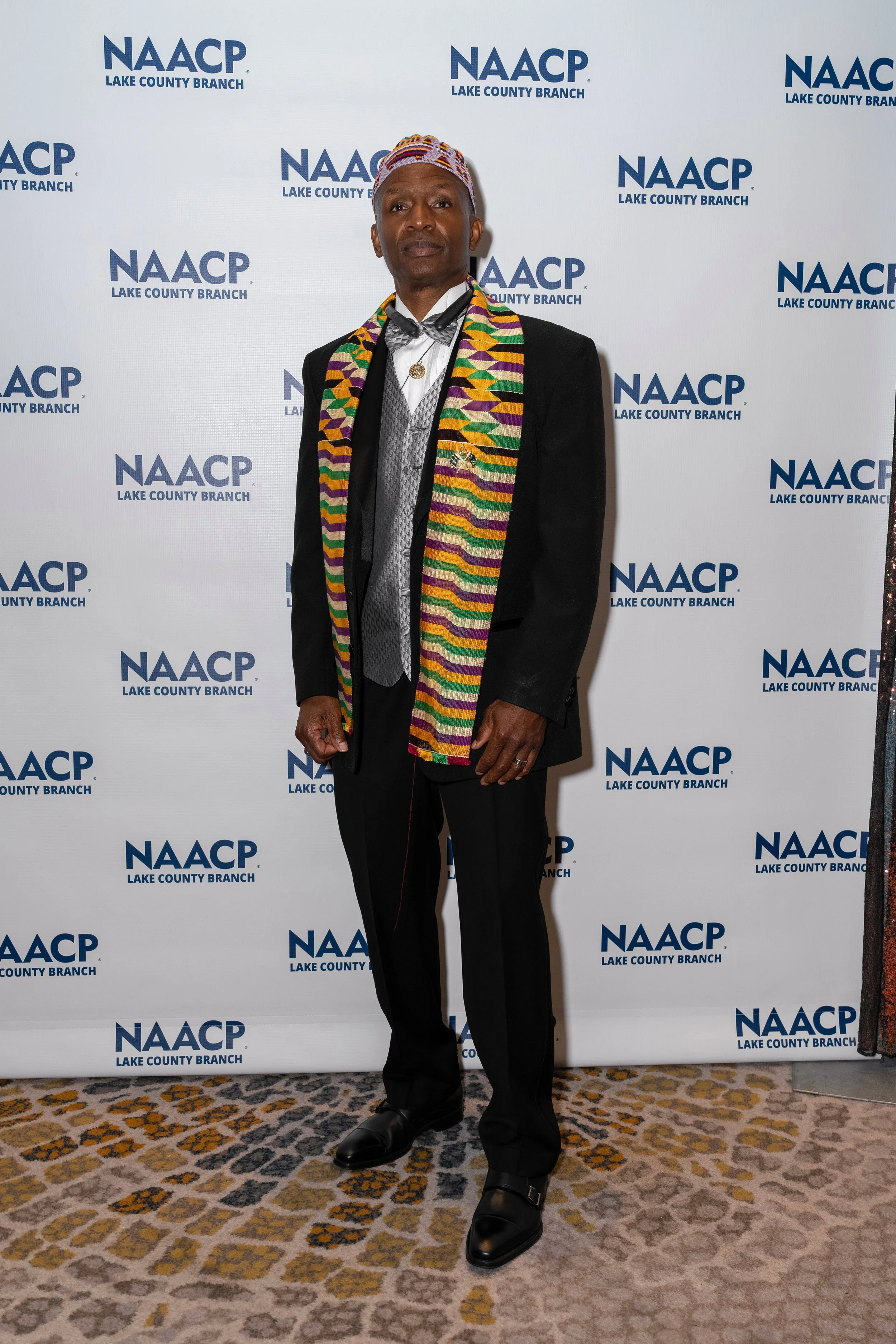 A man wearing a colorful patterned Kente cloth scarf, a traditional African hat, and formal attire standing in front of a white backdrop with the NAACP Lake County Branch logo.