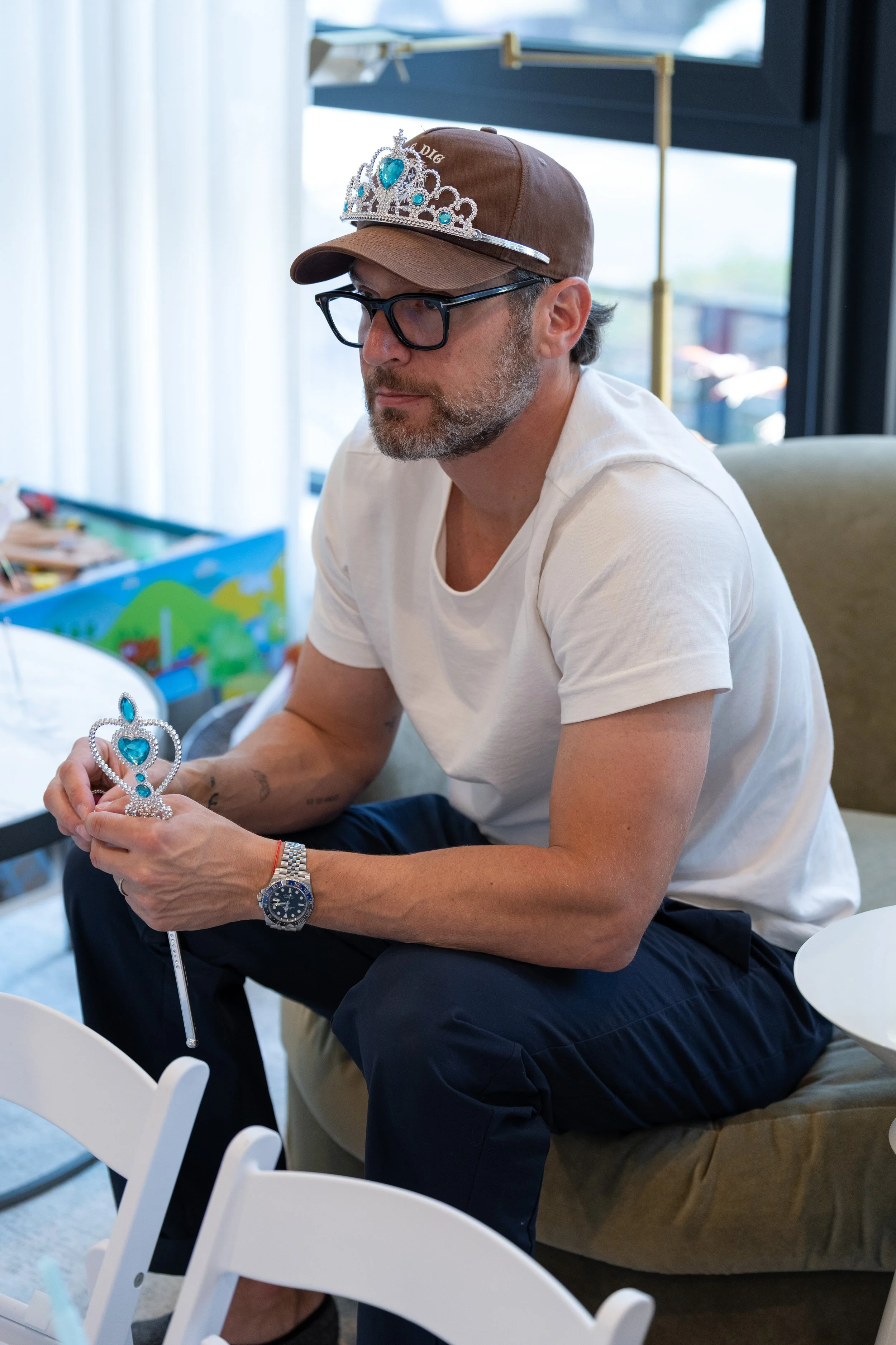 A man sitting on a beige couch holding a toy crown and wearing a tiara, with a baseball cap and glasses, in a room with large windows.