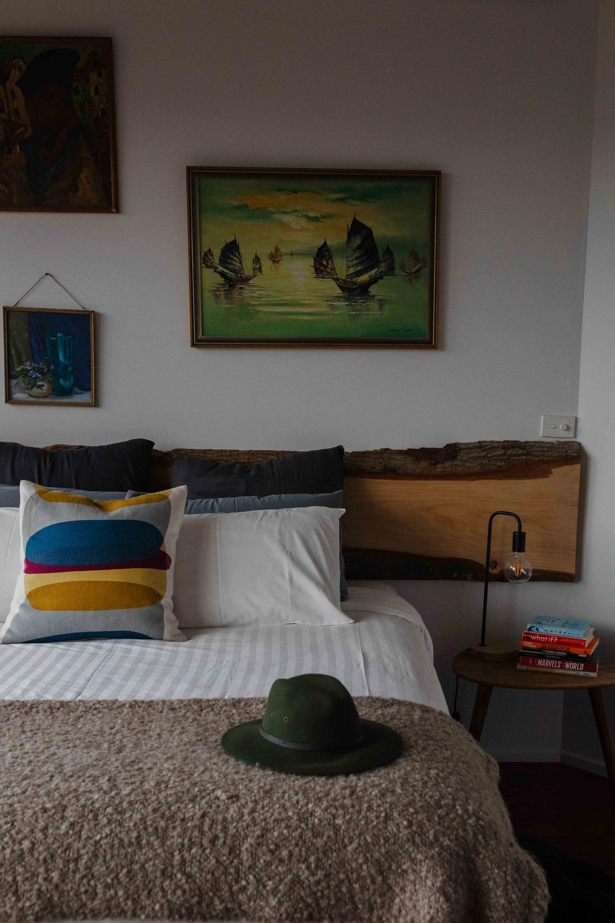 A bedroom with a bed having a white bedspread, colorful pillows, a wooden headboard, and a green hat on a brown blanket. There are framed paintings on the wall, a small round side table with a lamp, books, and a brown blanket.