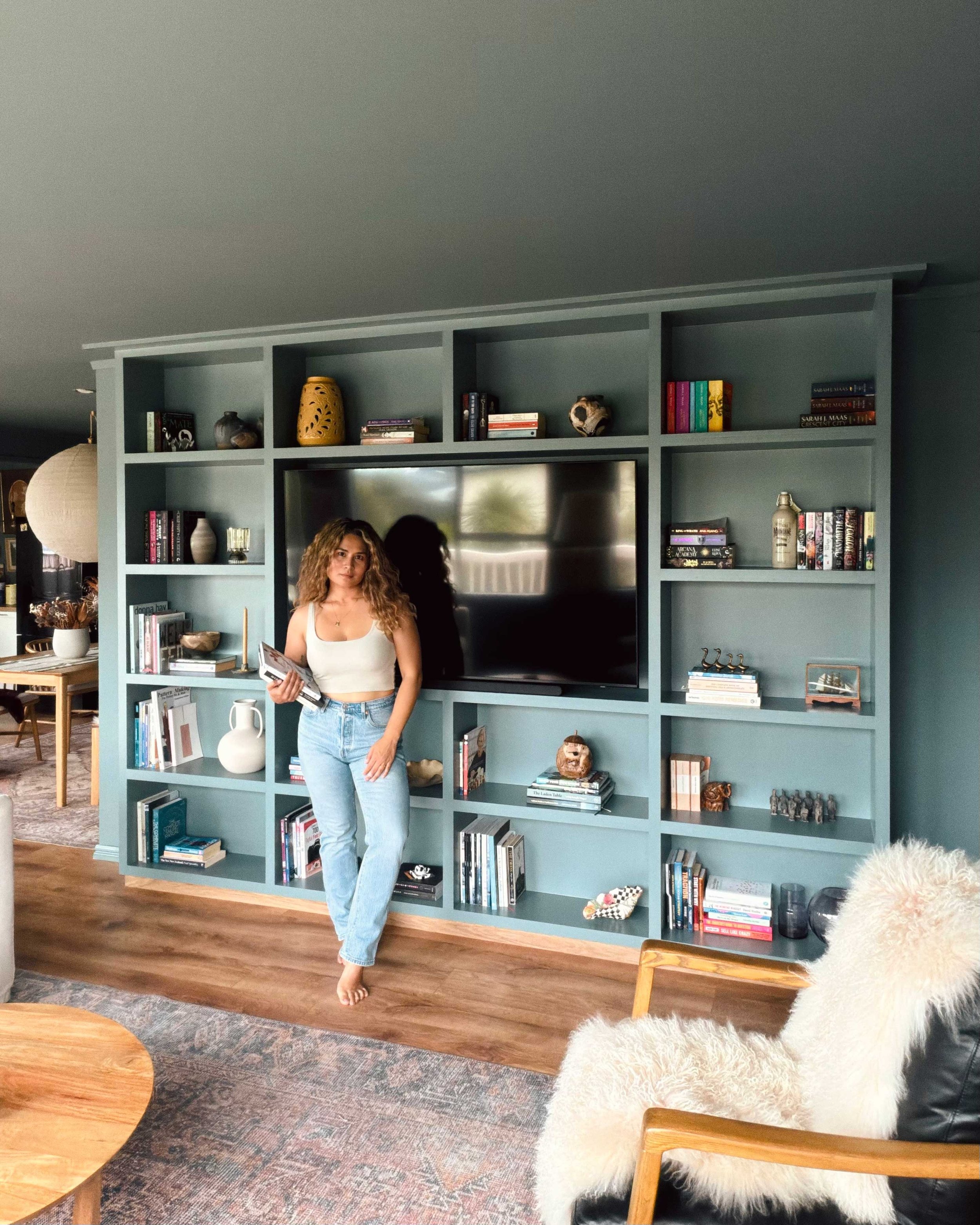 A woman with curly hair wearing a white tank top and jeans, standing barefoot in front of a teal bookshelf with a TV, books, and decorative items inside. The living room has wooden floors, a patterned rug, a wooden coffee table, and a cozy armchair with a fluffy white throw.