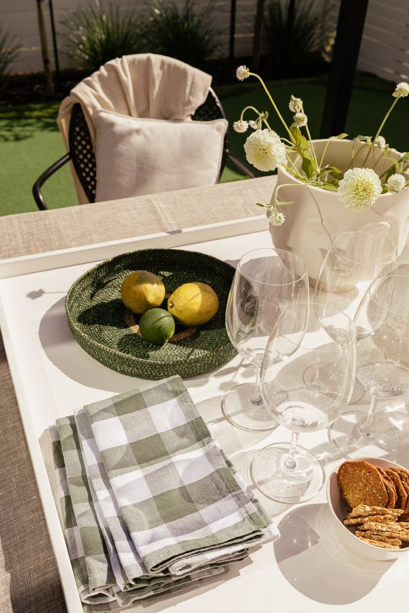 An outdoor dining table with a white tray, a green and white checked napkin, a bowl of cookies, clear wine glasses, a green fruit bowl with lemons and limes, and a vase with white flowers, set for evening dining.