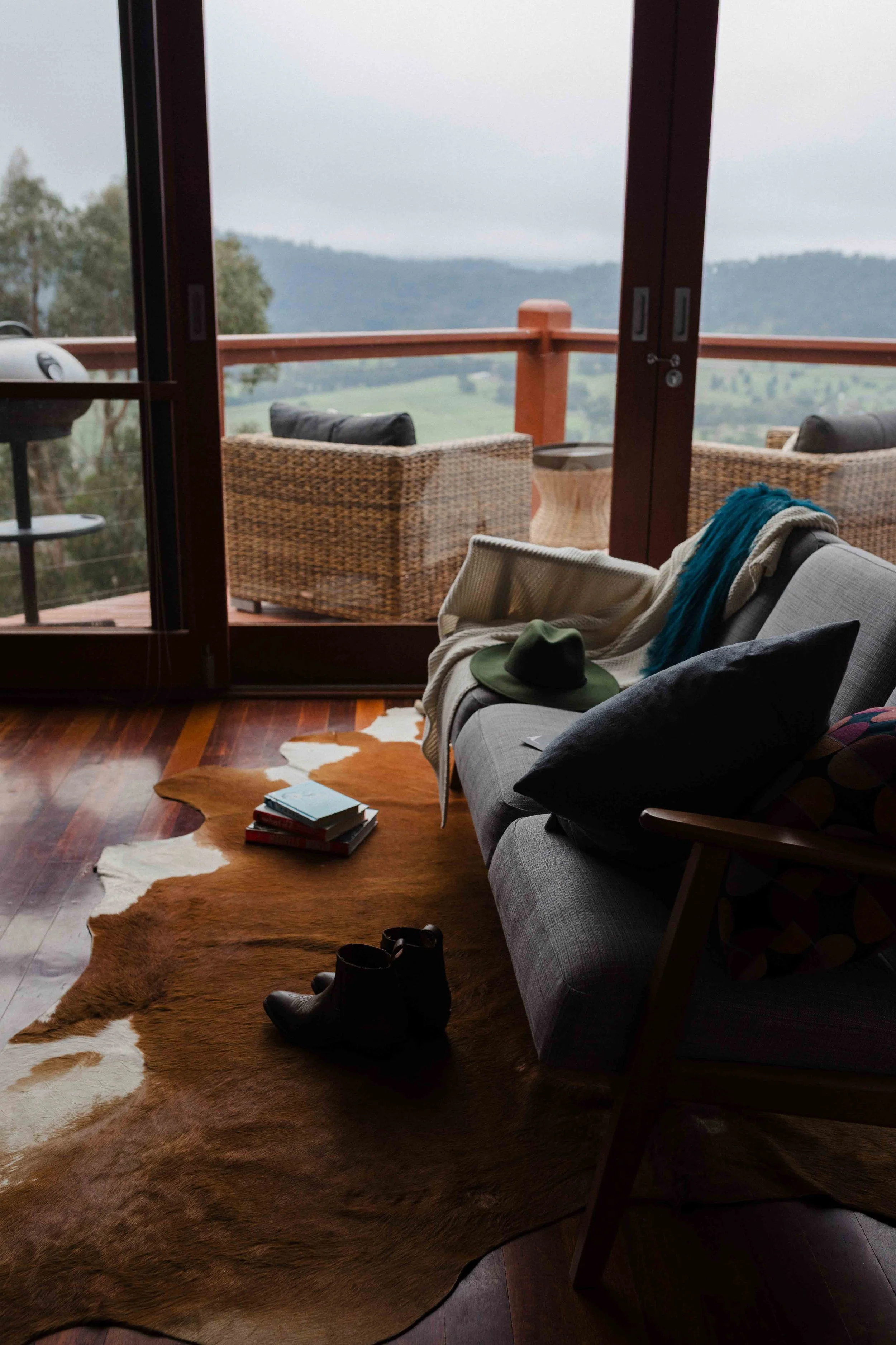 Living room with a gray couch, black pillow, books, a hat, and shoes on a cowhide rug, overlooking a wooded landscape through glass doors.