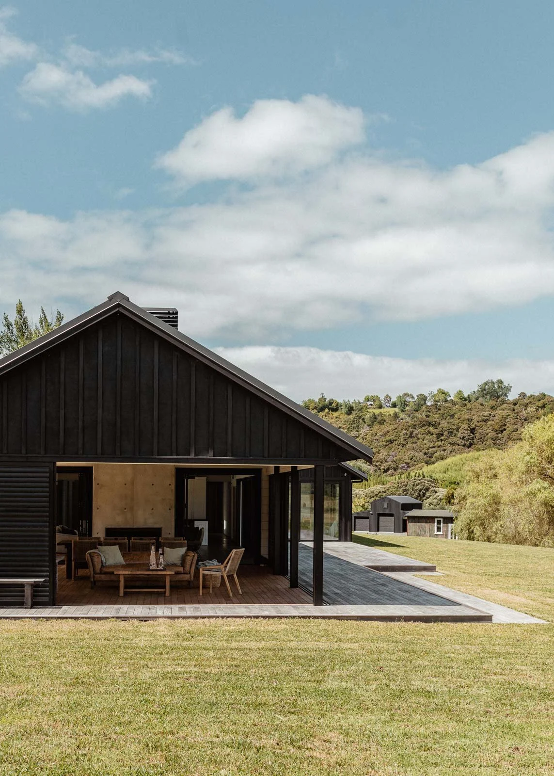 Modern black house with large glass windows and a covered porch, surrounded by green lawn and hills under a partly cloudy sky.
