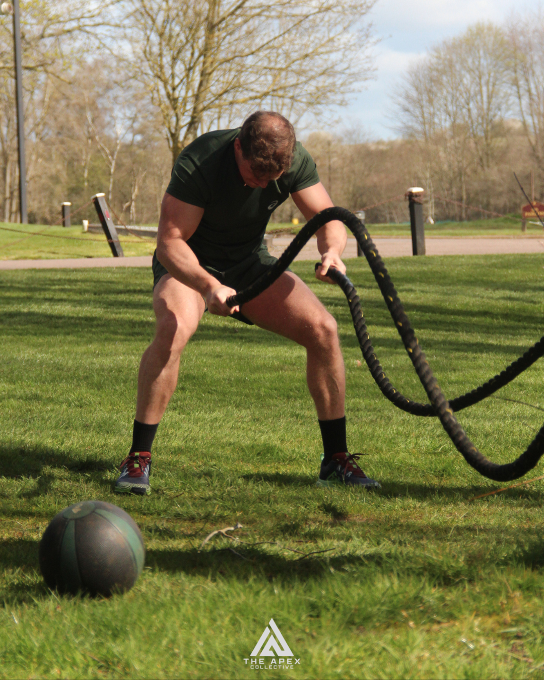 A man in athletic clothing engaging in battle rope exercises on a grassy field with trees and a parking lot in the background.