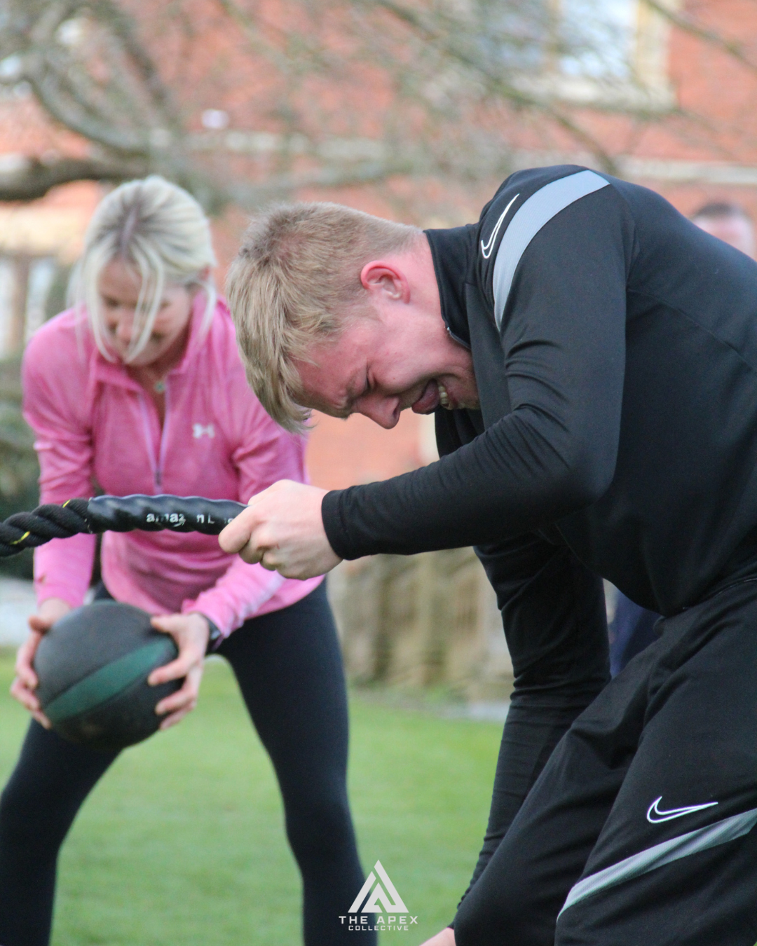 Two people, a man and a woman, engaged in outdoor fitness training, lifting a weighted ball and pulling a battle rope with an outdoor background.