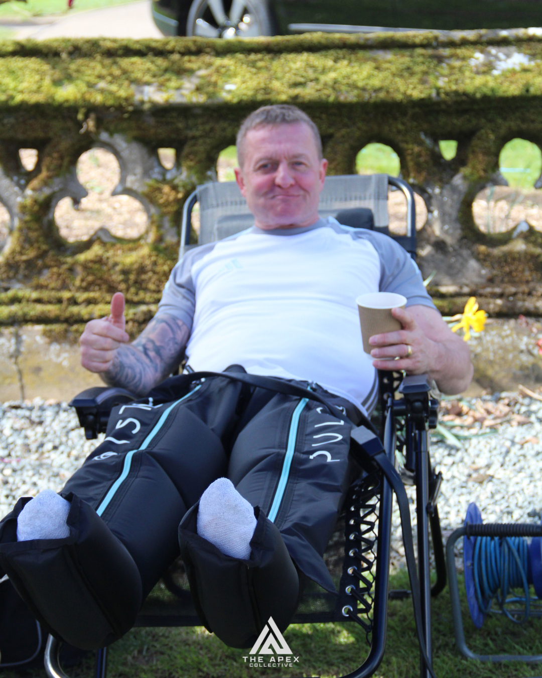 A man in a wheelchair outdoors, holding a paper cup and giving a thumbs up, with a moss-covered railing and greenery in the background.