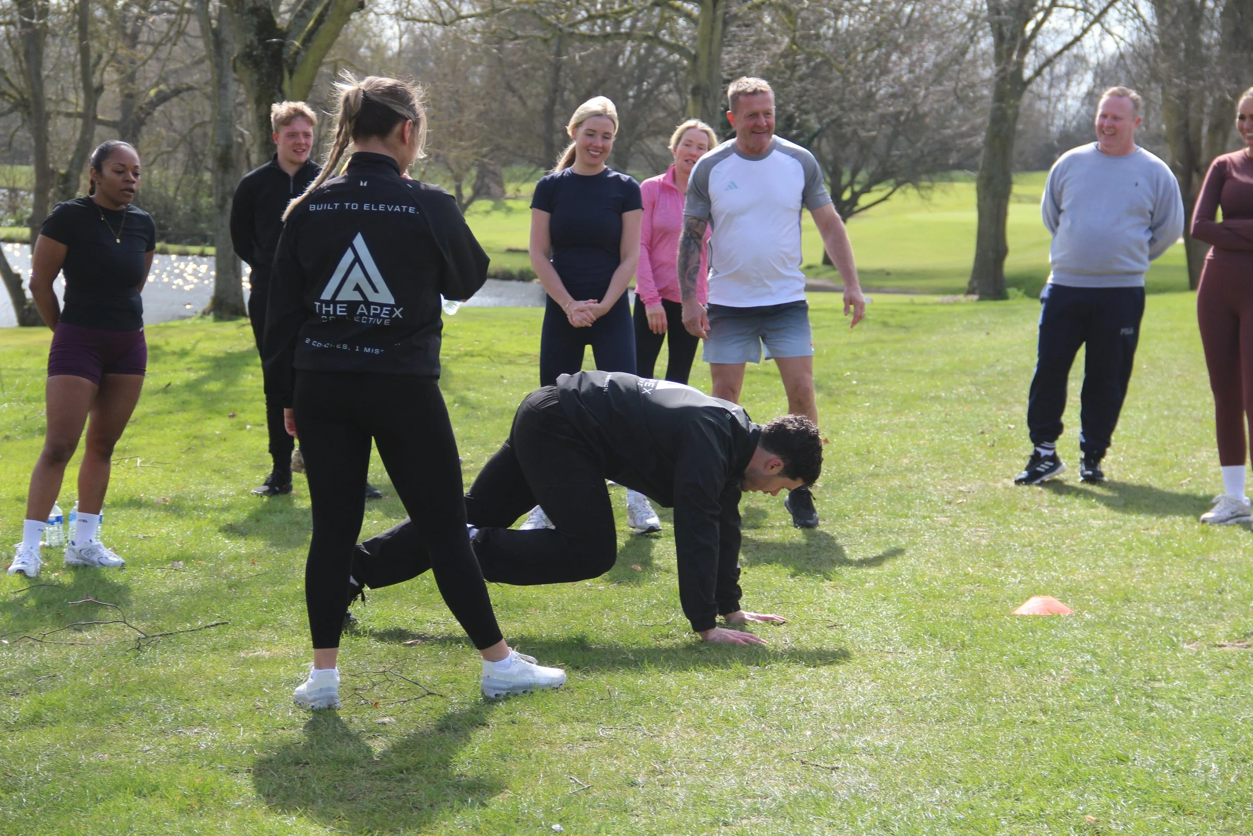 A group of people outdoors on a grassy area with trees, standing and watching someone perform push-ups. The group includes both men and women dressed in casual and athletic clothing, smiling and engaged in a fitness or training session.