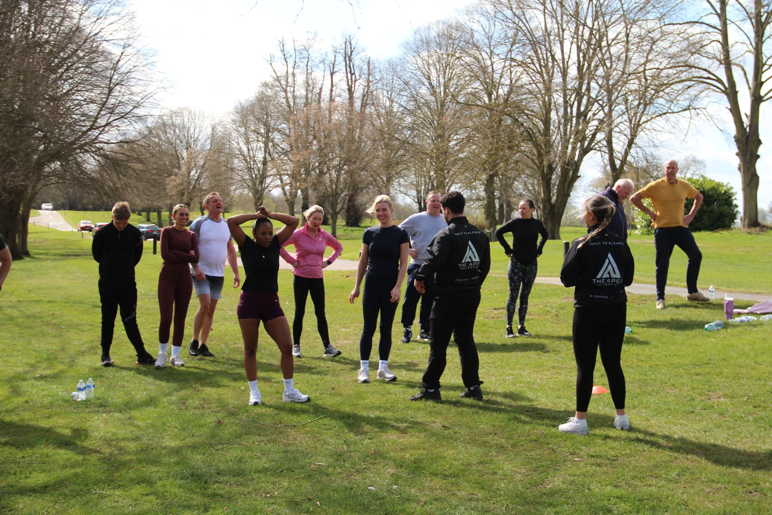 Group of people gathered outdoors in a park on a cloudy day, participating in a fitness or exercise class, with some standing and others stretching or talking, surrounded by trees and parked cars.