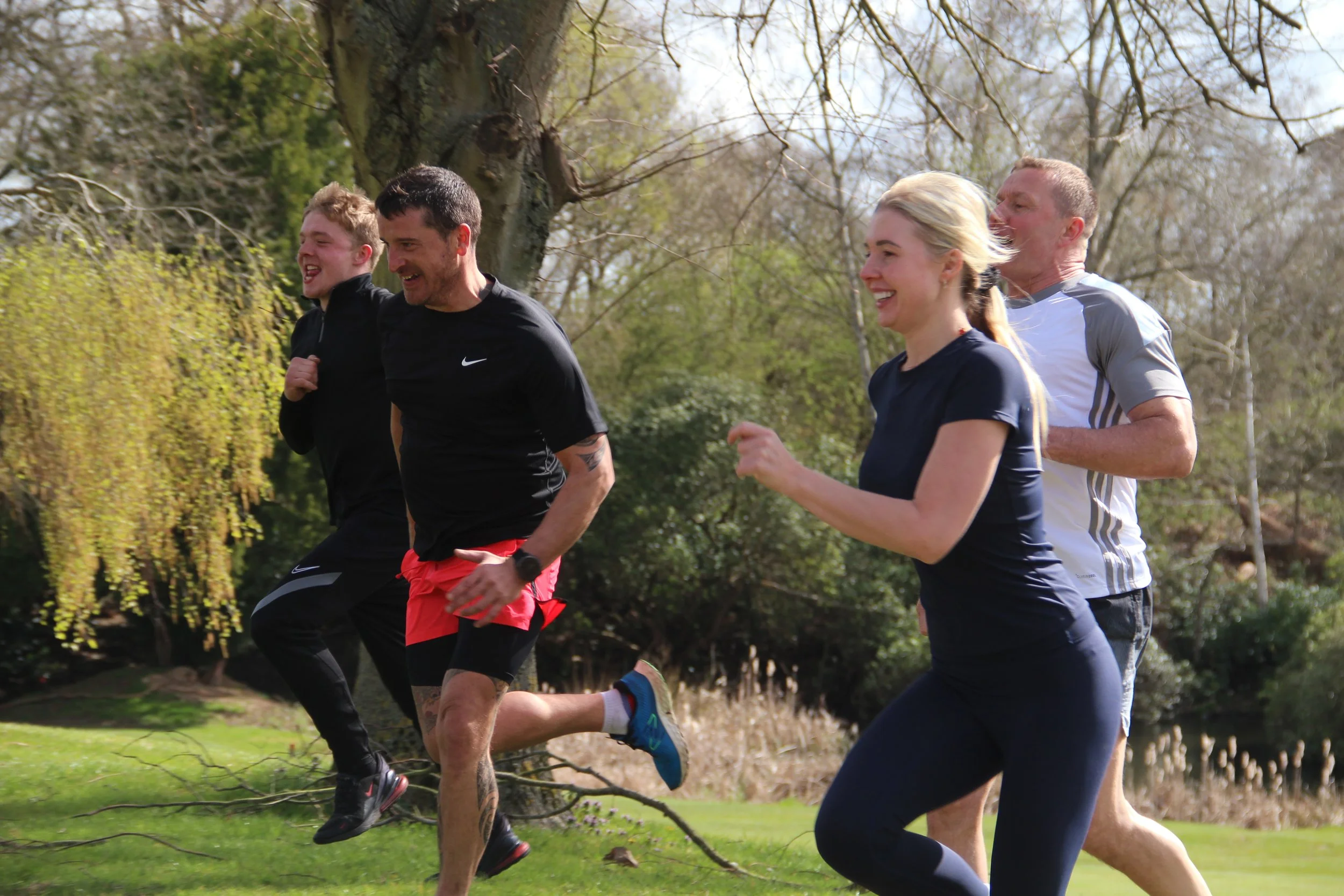 Four people, two men and two women, jogging outdoors in a park during daytime, with trees and greenery in the background.