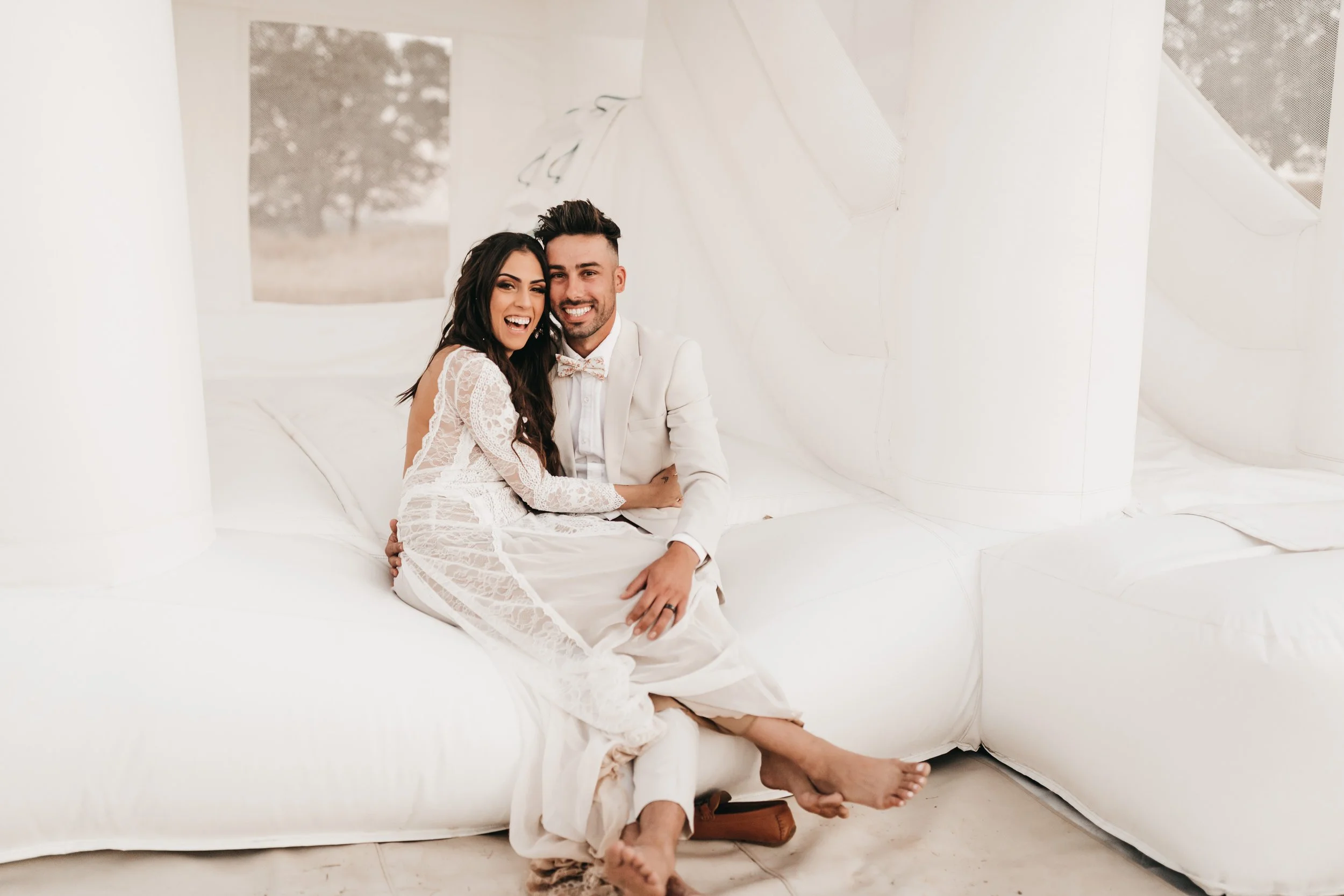 A happy couple, dressed in wedding attire, sitting on a white inflatable jump house outdoors, smiling and embracing each other.