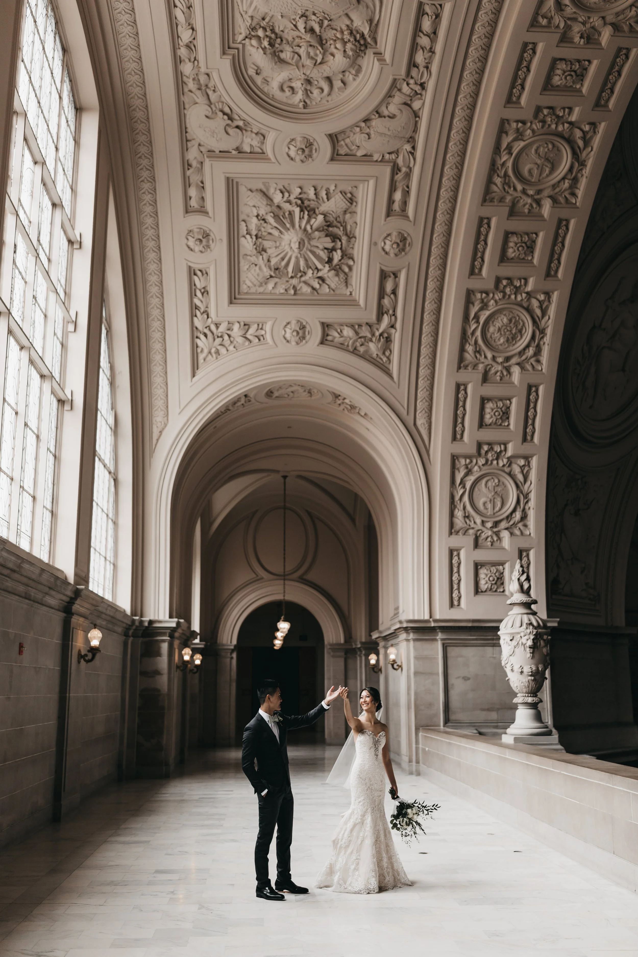 A bride and groom dancing in a grand, ornate hall with high ceilings and decorative moldings.
