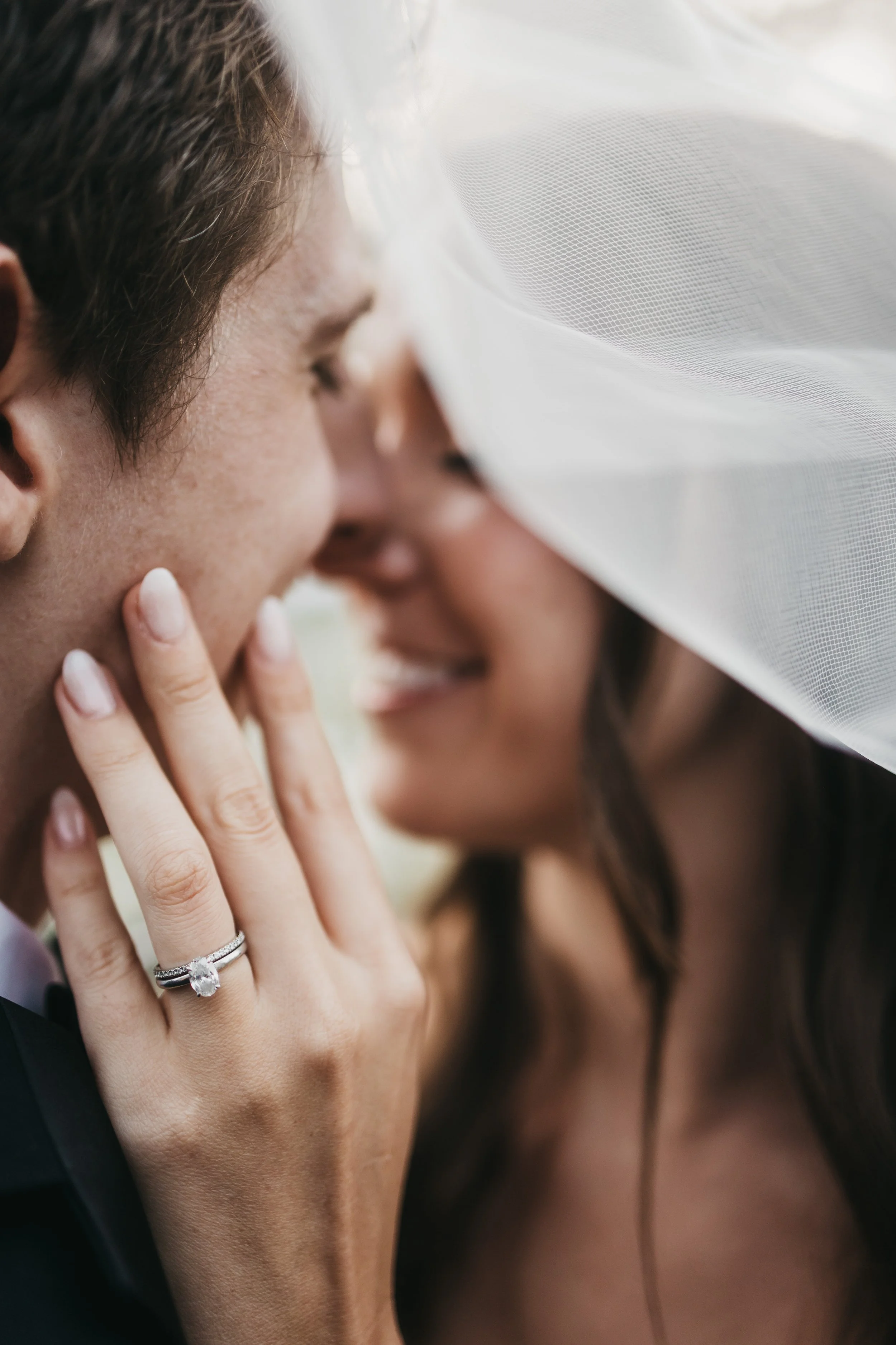 Close-up of a couple about to kiss, with a woman's hand gently touching the man's face, showing an engagement ring. The woman is wearing a veil.