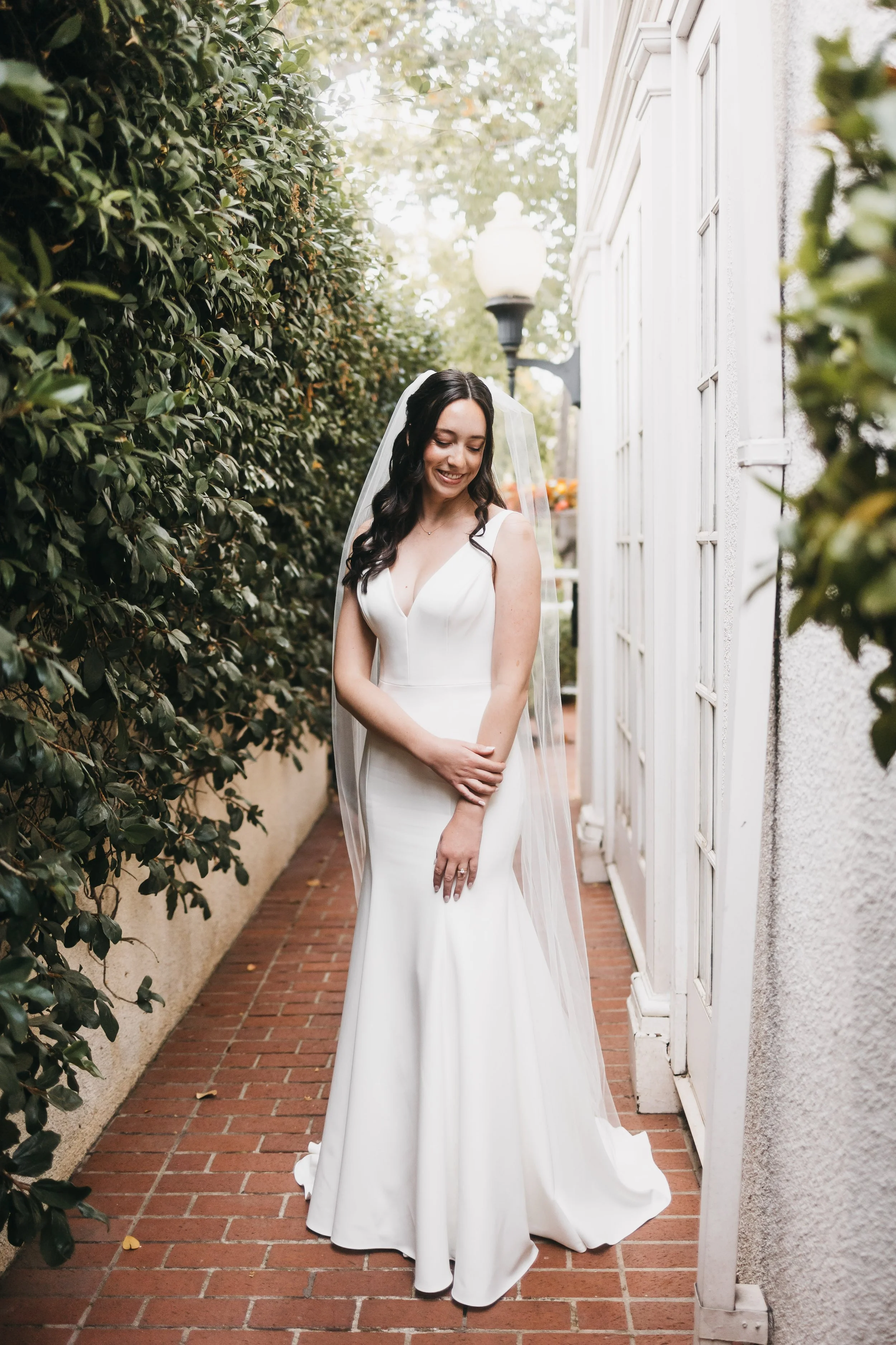 A bride in a white wedding gown and veil stands on a brick pathway next to a white building wall, smiling and looking down with her hands crossed in front of her.