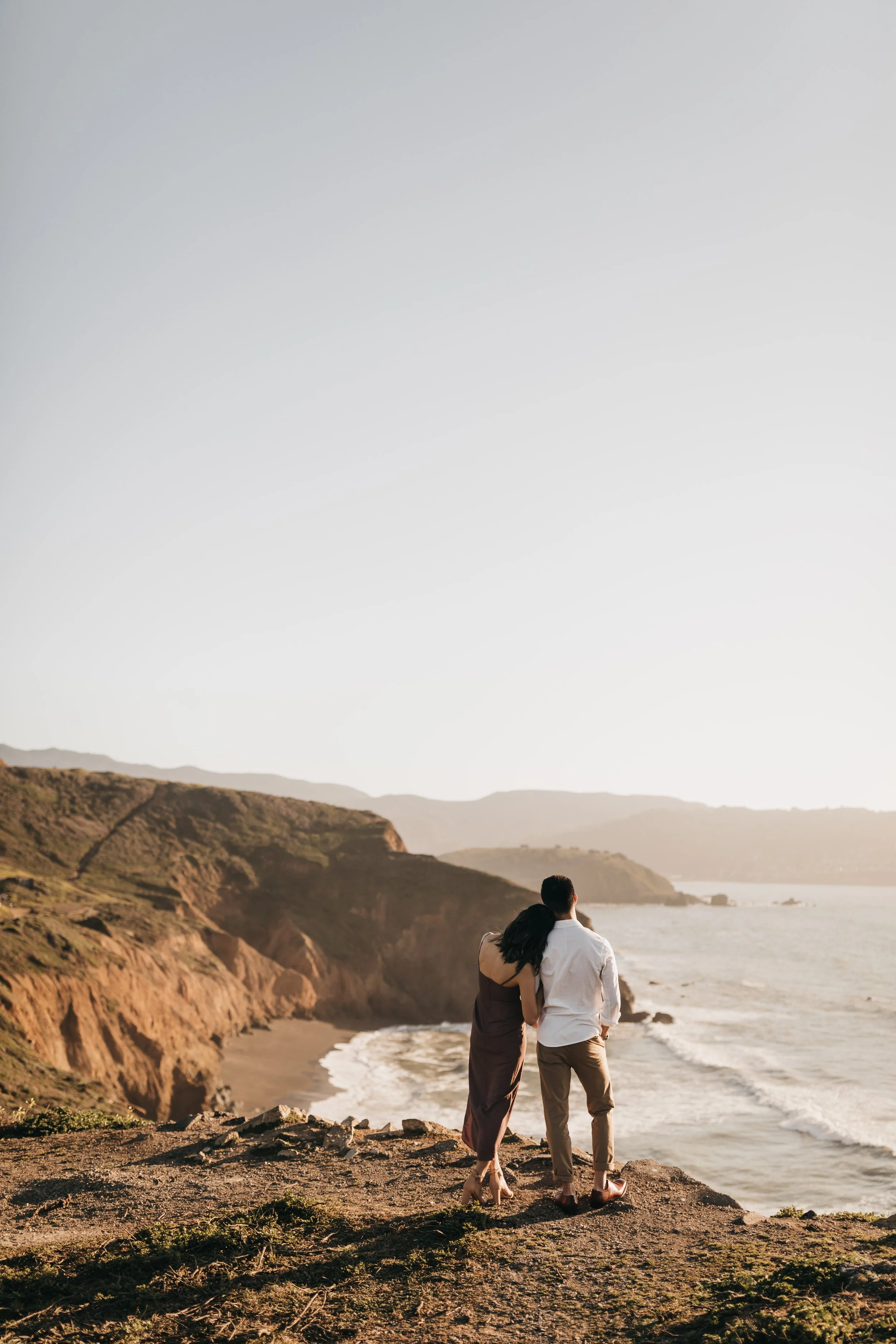 A couple standing on a cliffside overlooking the ocean during sunset, embracing each other.