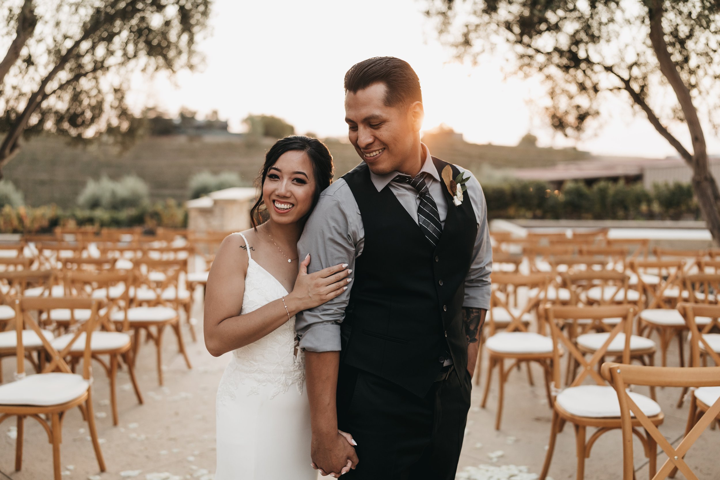 A wedding couple smiling and holding hands outdoors with rows of chairs and trees in the background during sunset.