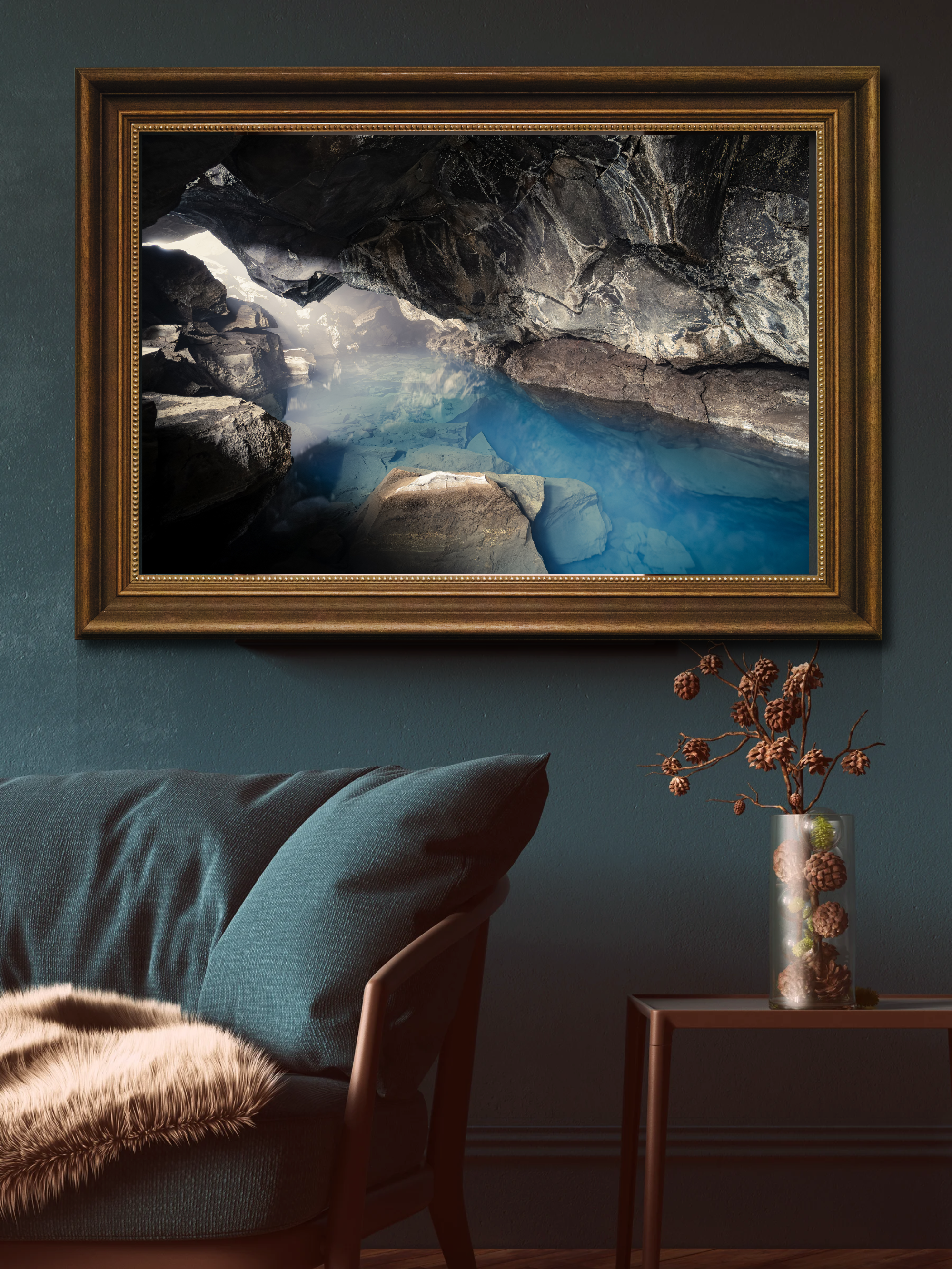 A framed picture of a cave with water and rocks hanging on a green wall, with a teal chair with a fur throw and a side table with a vase of decorative pinecones and branches.