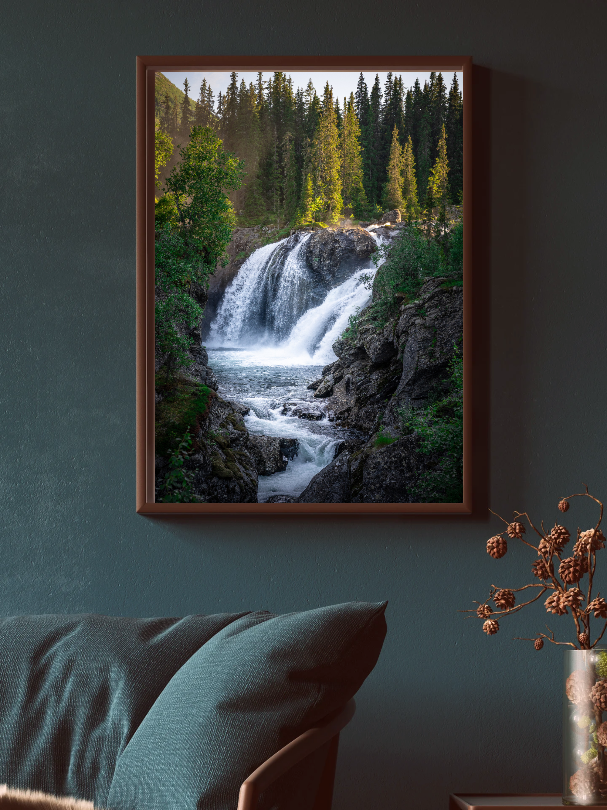 Framed picture of a waterfall with trees on either side, located on a wall in a room with a green panel. A sofa with a pillow and a decorative vase with dried plants are visible in the room.