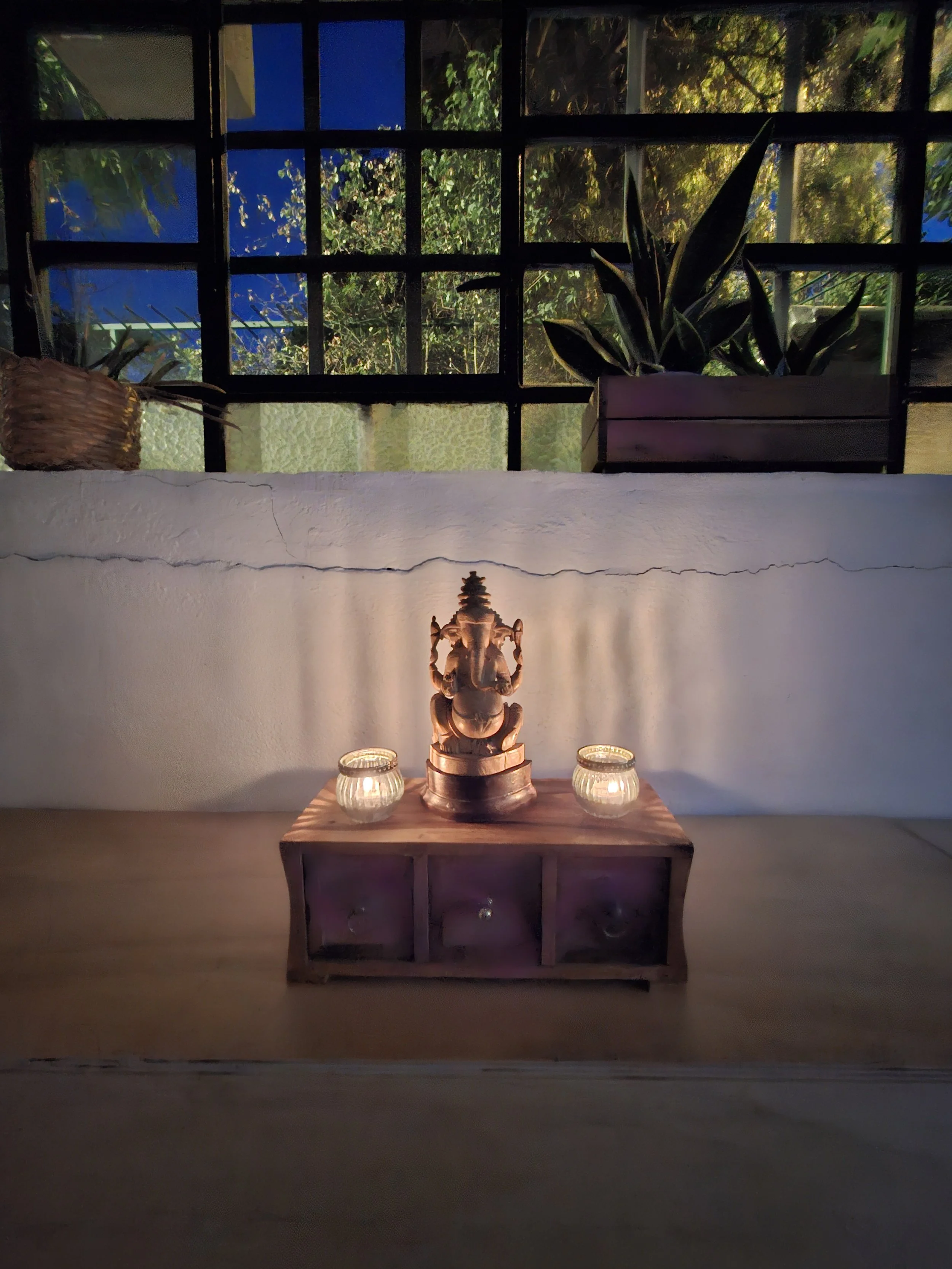 Estatua de Ganesha en una mesa de madera con dos velas a su lado, en un ambiente interior con ventanas y plantas en el fondo.