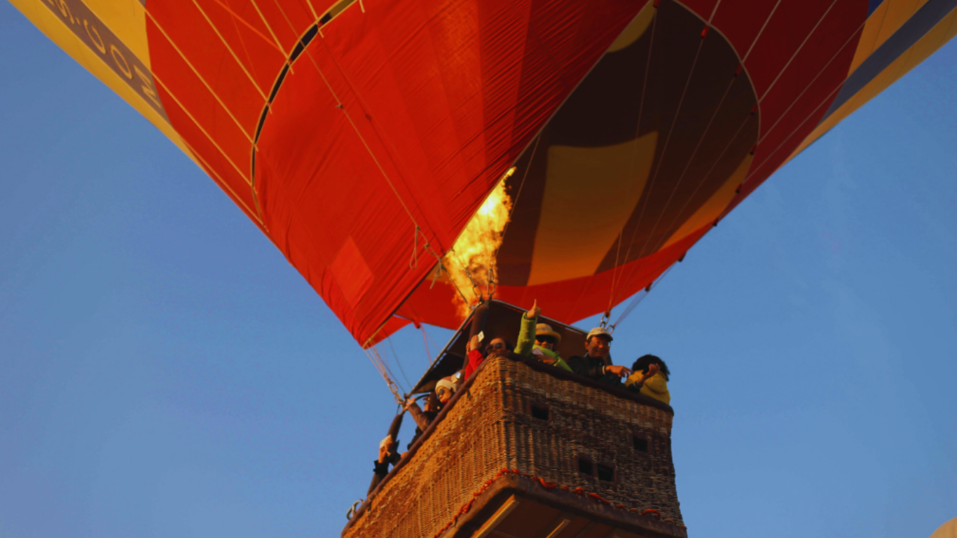 Montgolfière en vol avec un groupe de personnes dans la nacelle, déployant une flamme pour faire chauffer l'air dans le ballon, contre un ciel clair.
