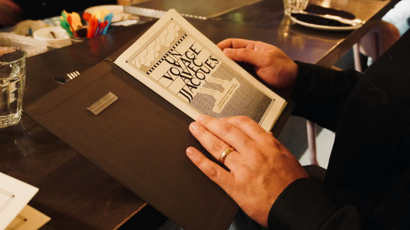 Une personne tient un menu de restaurant intitulé "Un voyage à Jacouës" dans une main, avec une table en bois sombre devant elle, une verre d'eau à gauche, et des couverts en arrière-plan.