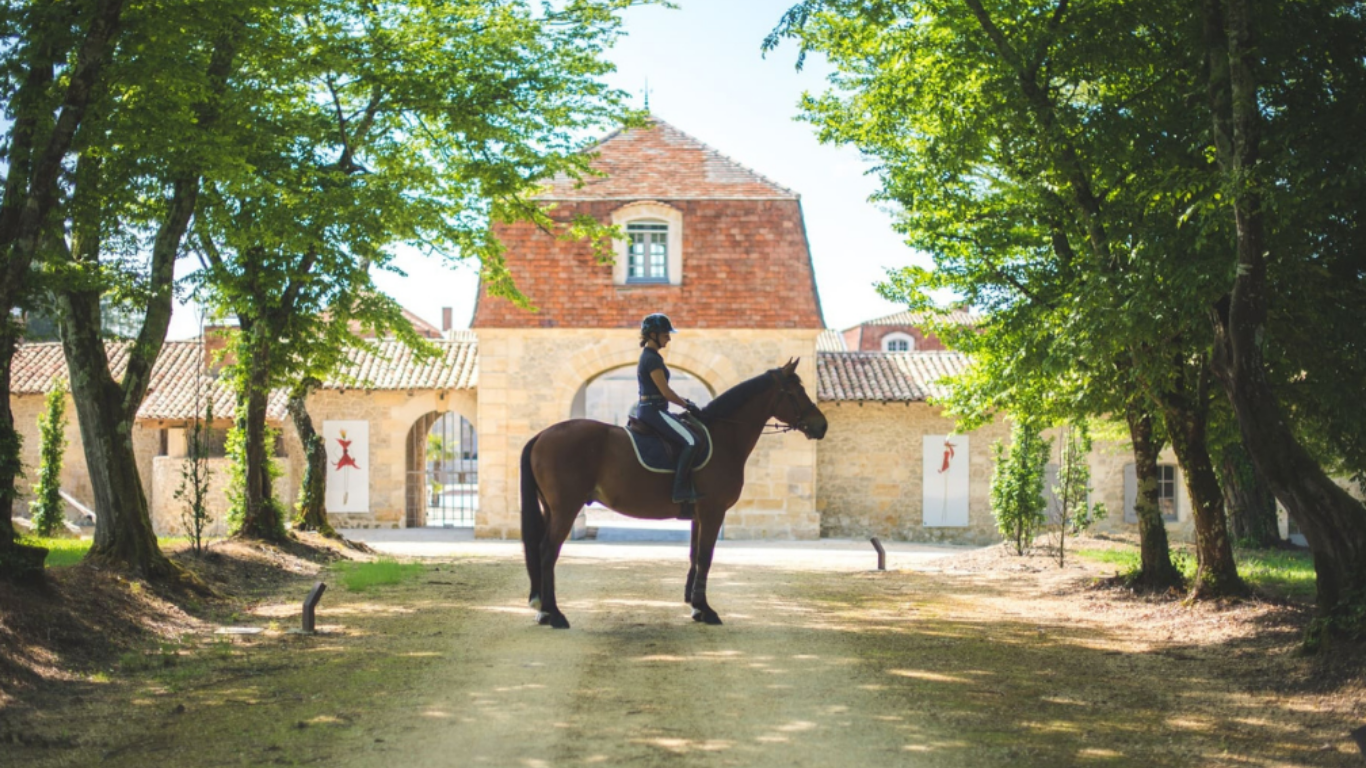 Une personne à cheval, portant un casque, devant un bâtiment en pierre avec un grand arbre à chaque côté, dans un environnement en plein air.