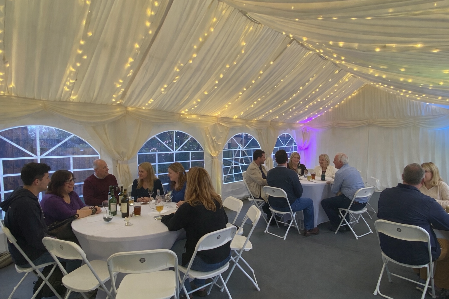 People sitting at round tables inside a decorated white tent with string lights and windows during a social gathering or celebration.