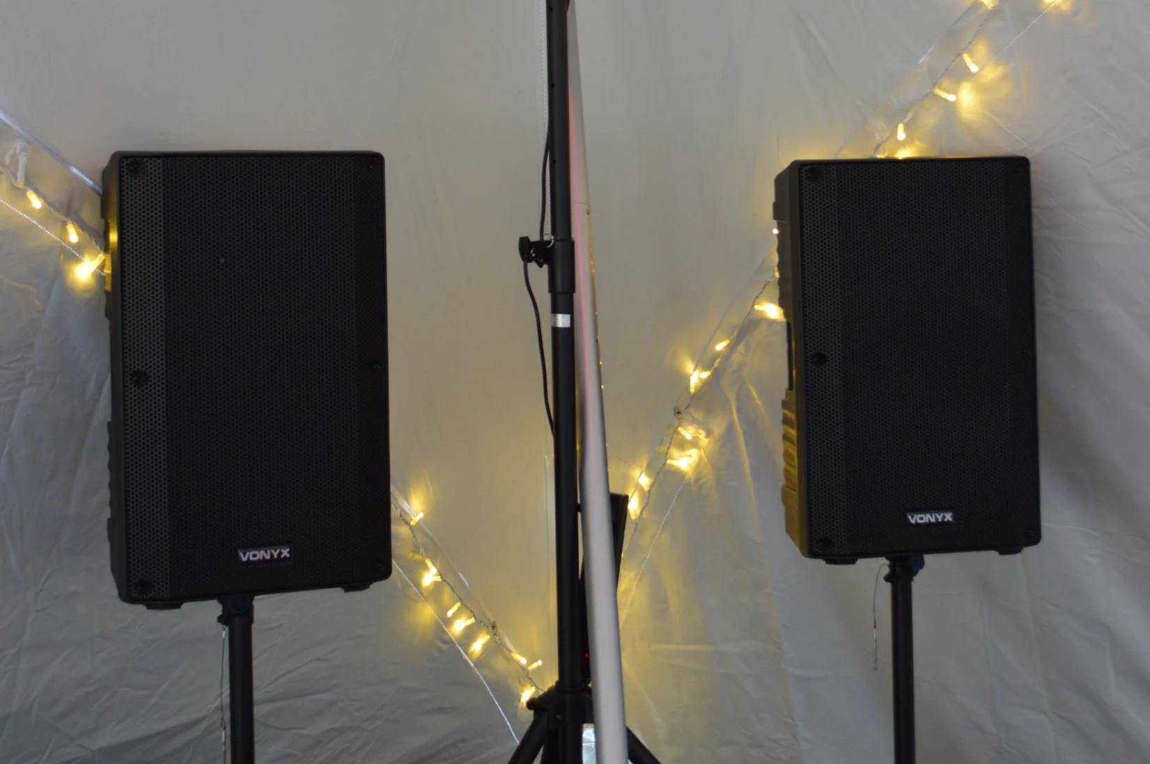 DJ equipment with two large speakers, lighting fixtures mounted on a stand, and string lights with yellow and white bulbs hanging against a white backdrop.