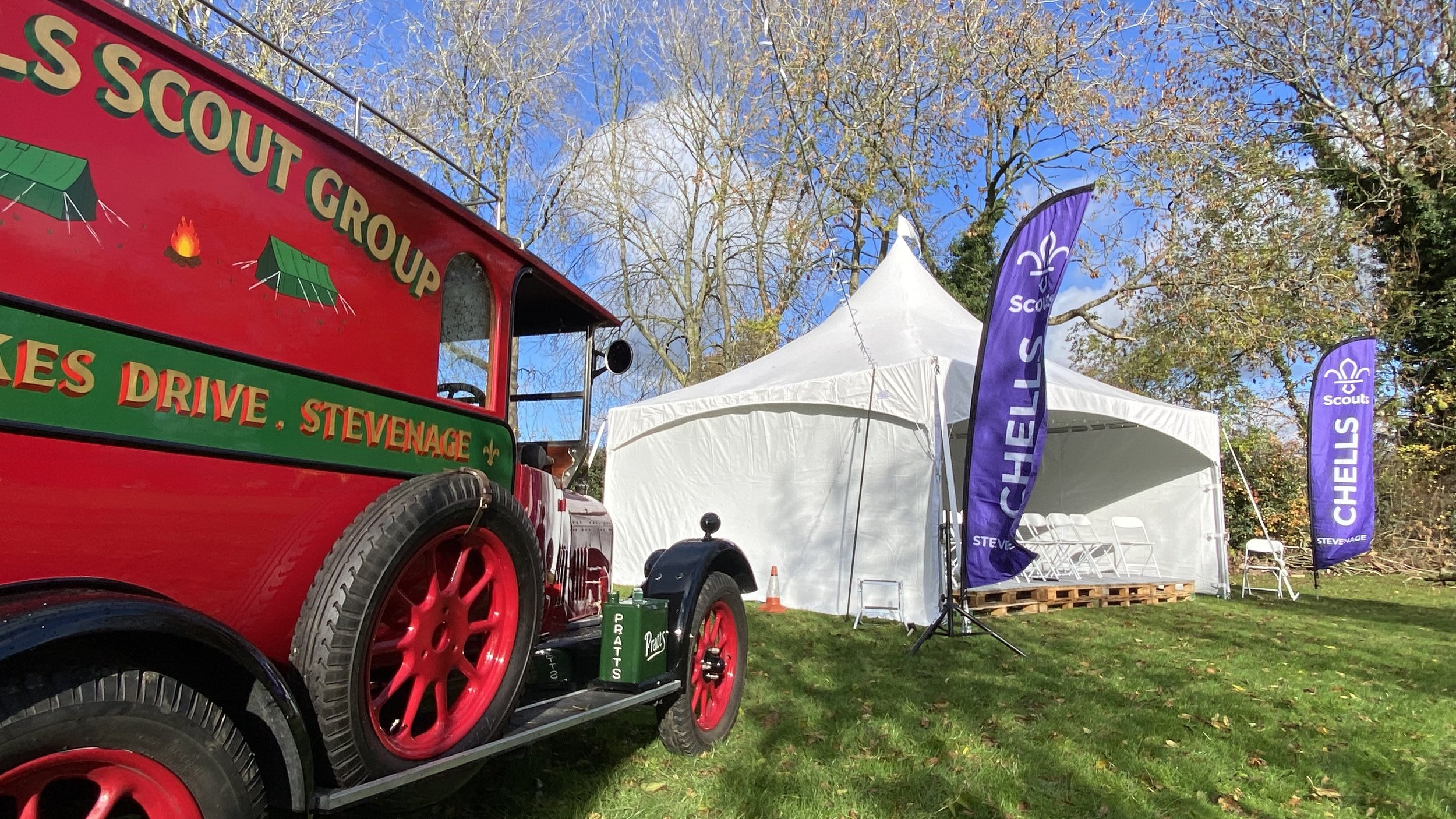 A vintage red Scout truck with green lettering and red wheels parked on grass outside a white tent with Scout flags, trees with fall foliage in the background, and a blue sky.