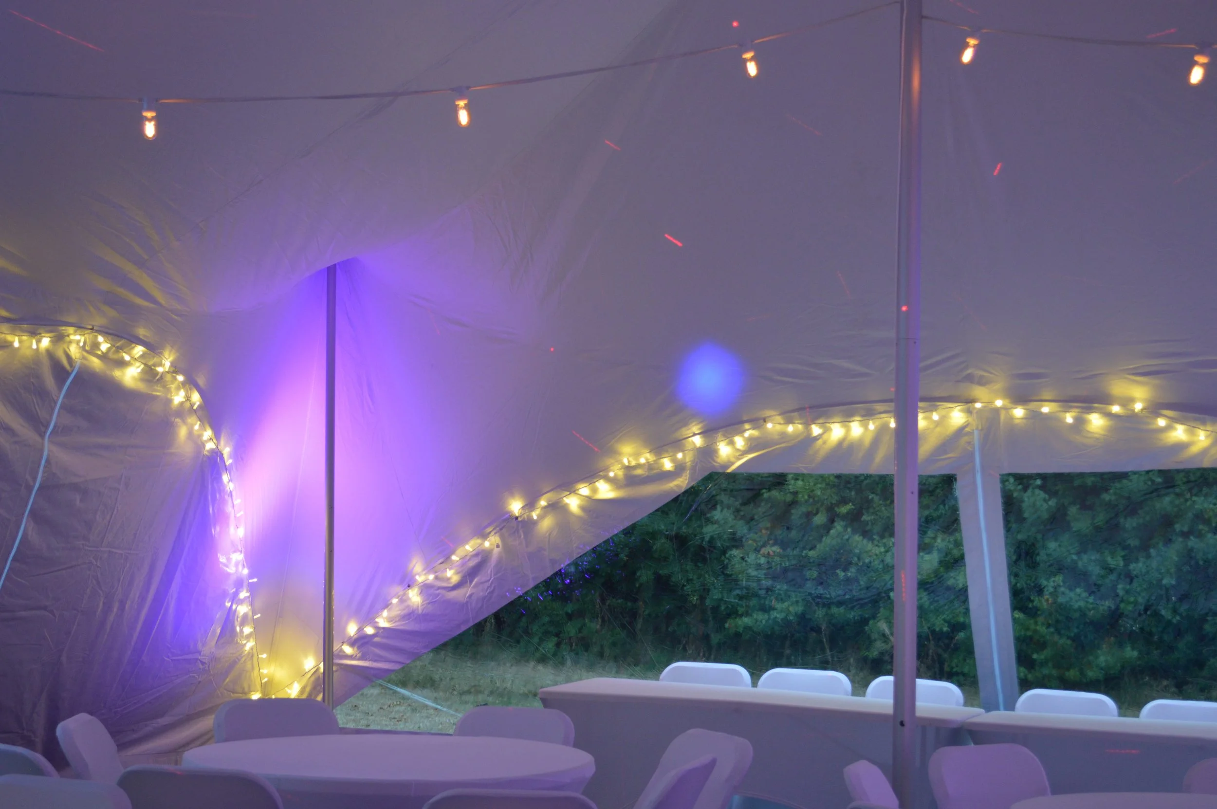 Interior of a tent decorated with string lights and purple and blue lighting, with white chairs and tables inside, overlooking greenery outside.