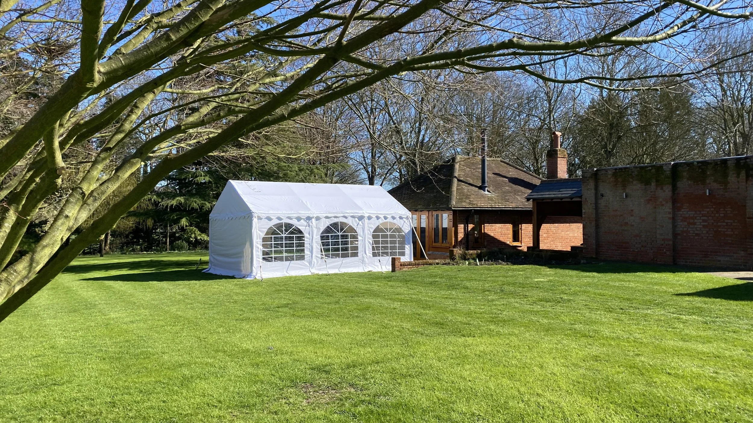 A white event tent set up on a lush green lawn, adjacent to a brick house with a chimney, under clear blue skies and leafless trees.