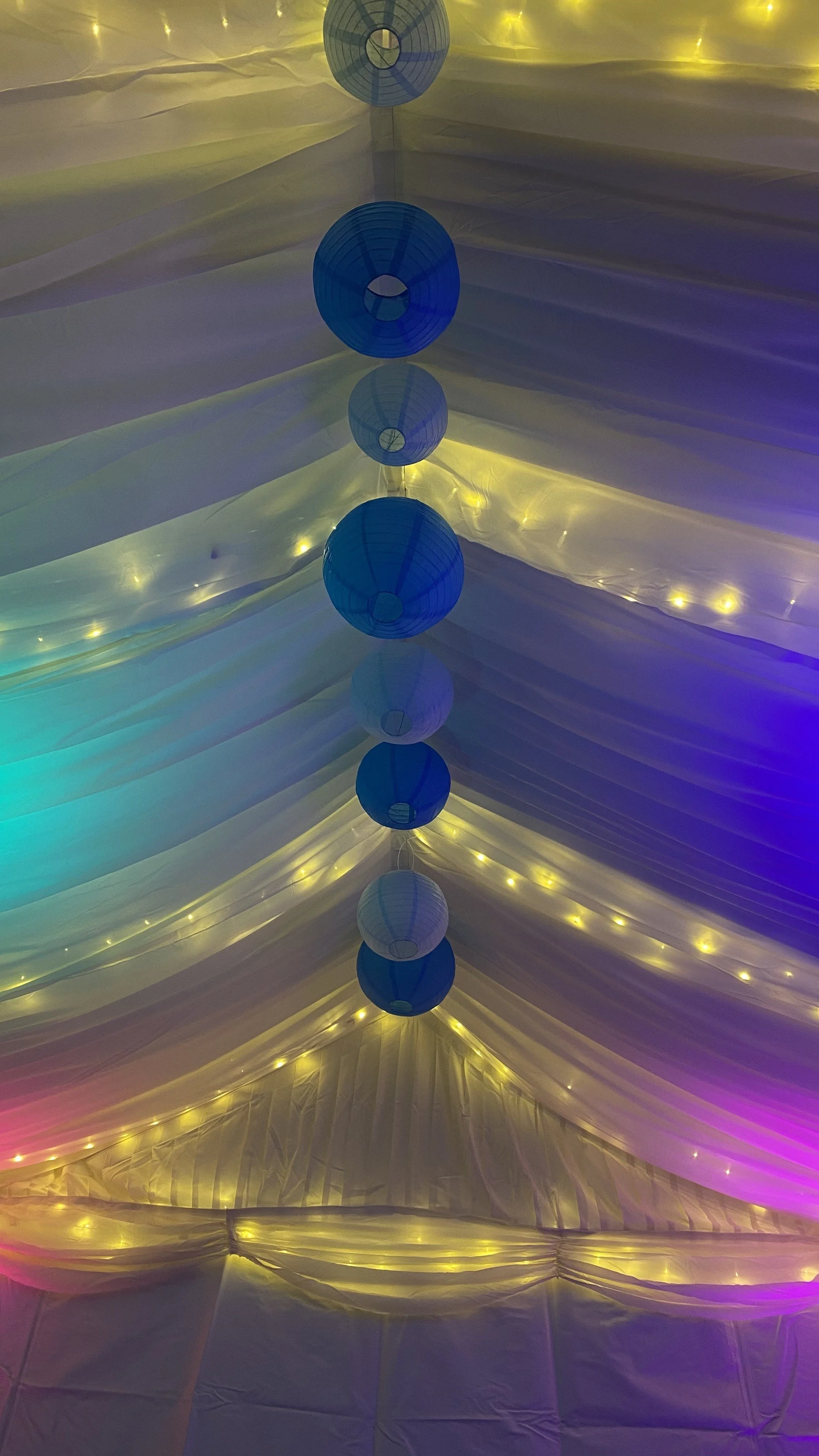 Looking up into the apex filled with blue paper shades. Warm white LED shining through the curtains and coloured uplighitng