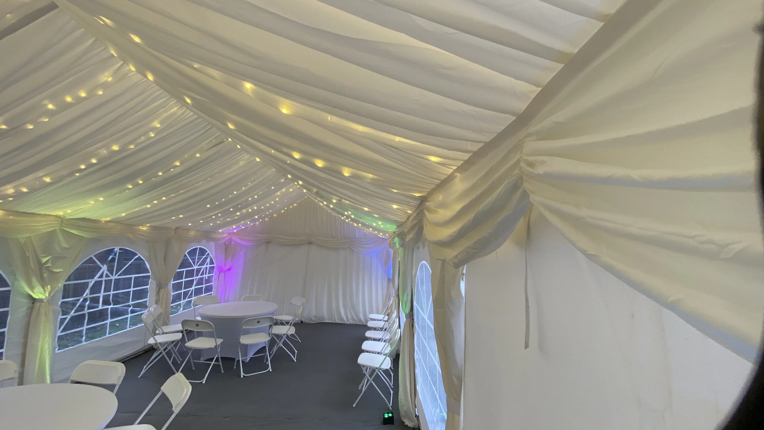 Interior of a decorated event tent with white drapery, string lights on the ceiling, and colorful uplighting along the sides. White round tables and chairs are arranged inside.