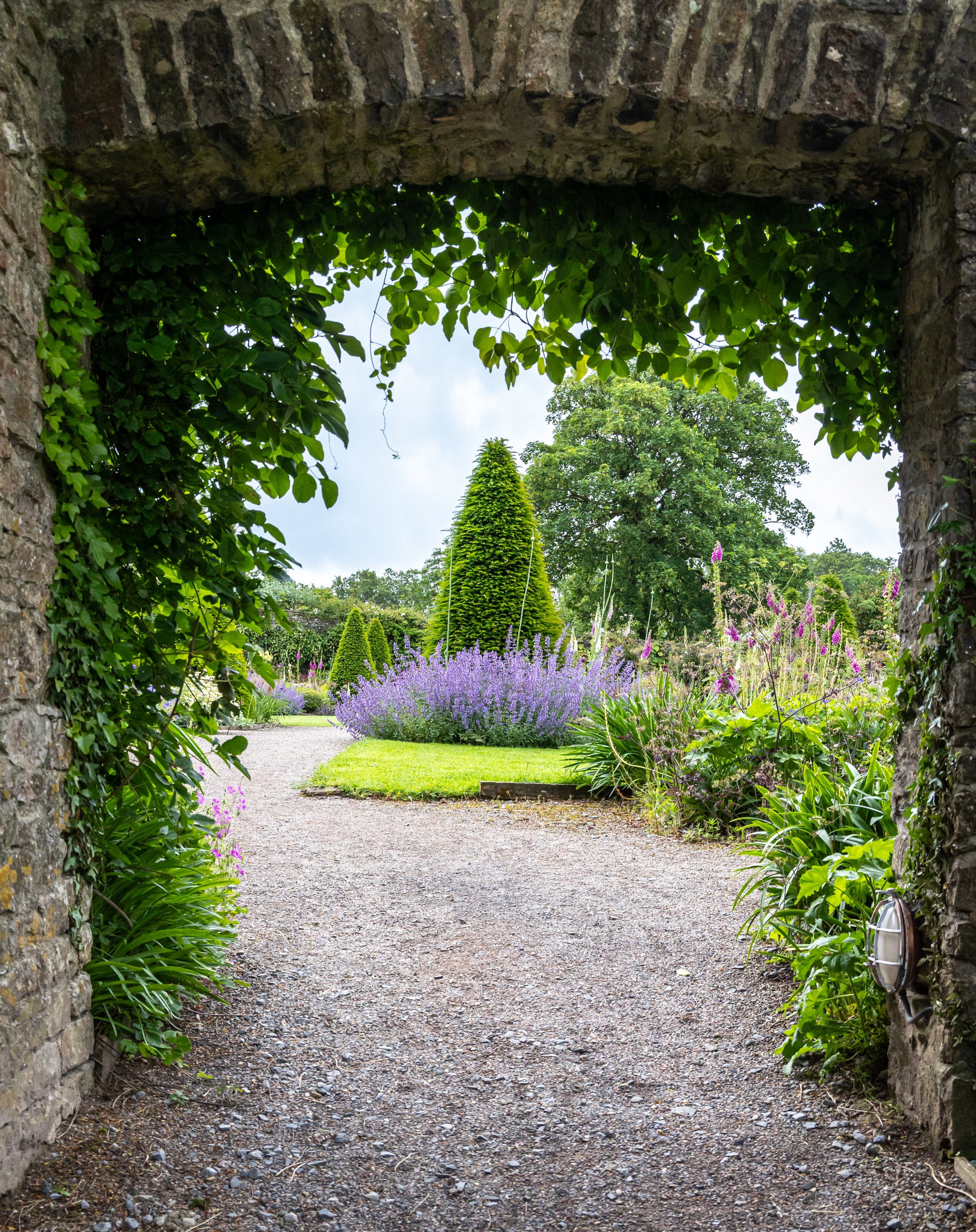 A view of a garden seen through an arched stone gateway with green foliage and blooming plants, including lavender and tall trees in the background.