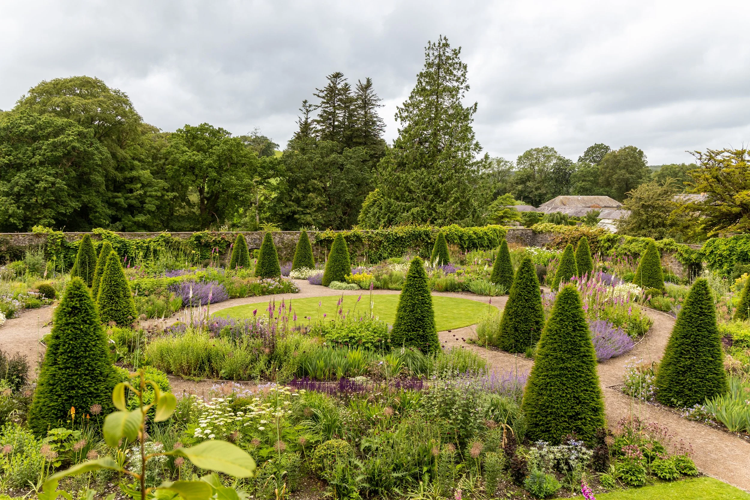 A lush botanical garden with a circular grassy area surrounded by pathways, colorful flowers, and cone-shaped shrubs, with a variety of trees in the background under a cloudy sky.