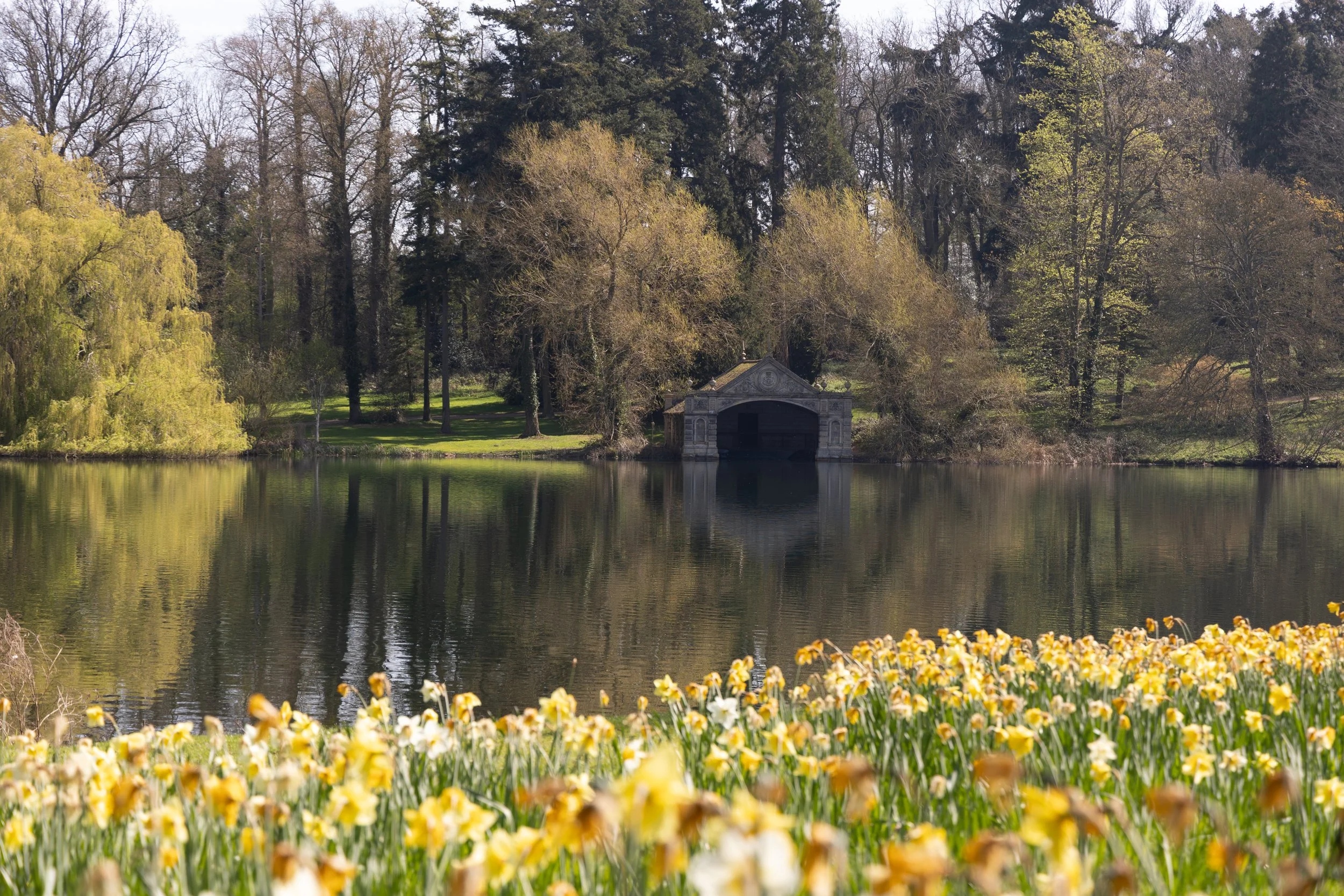 A serene park scene featuring a pond with a small stone boathouse, surrounded by trees, some with green leaves and others bare, with yellow flowers in the foreground.