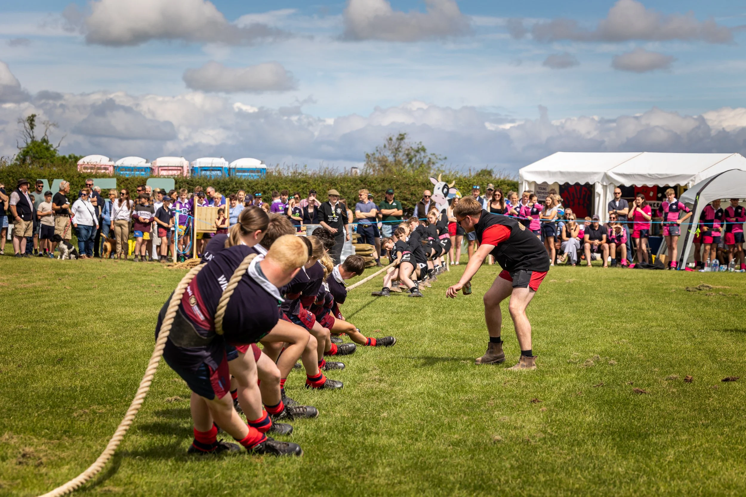 People participating in a tug-of-war game on a grassy field during an outdoor event, with onlookers watching and tents set up in the background under a partly cloudy sky.