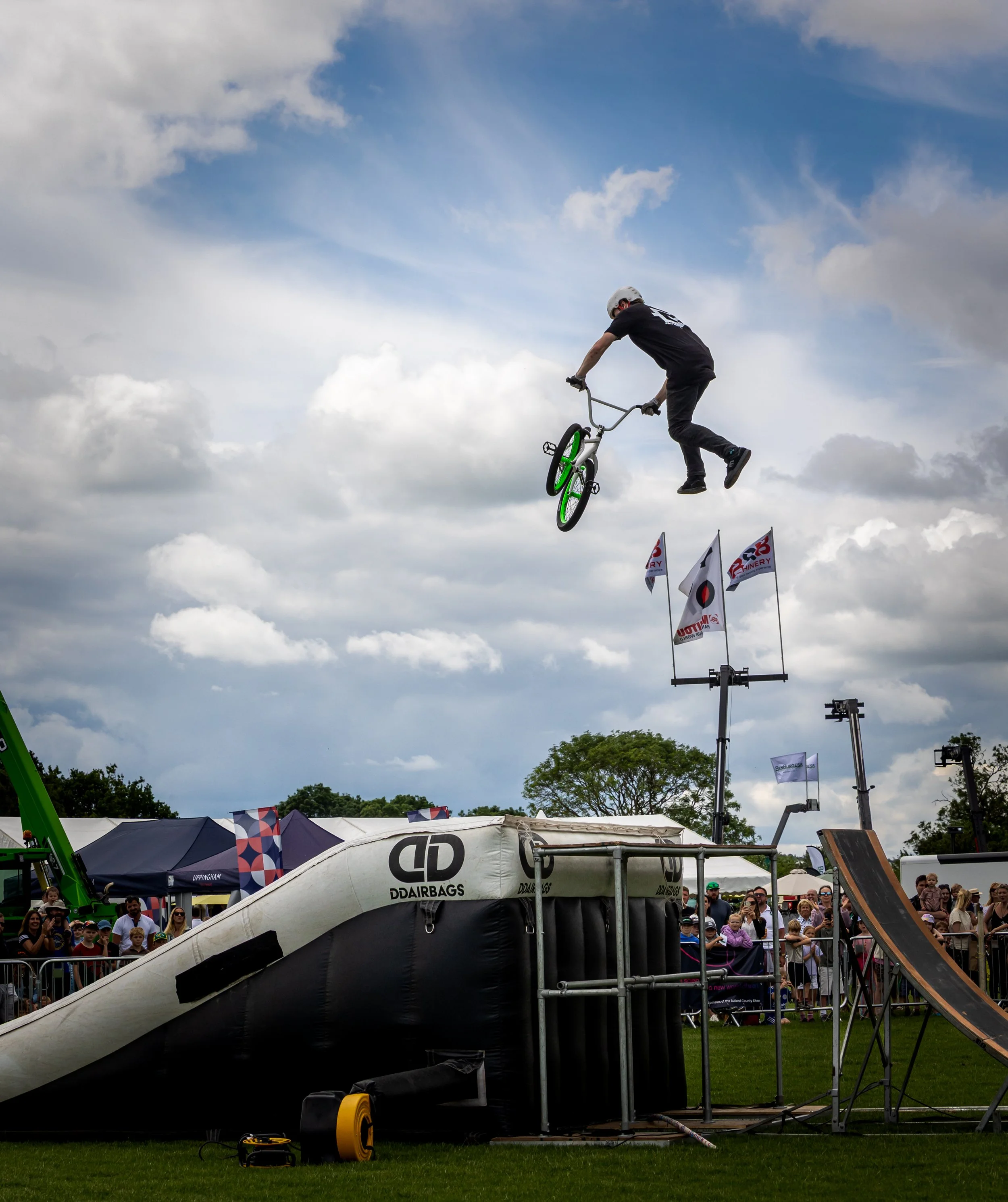 A person is mid-air, performing a trick on a BMX bike at an outdoor event with a crowd watching. The sky is cloudy.
