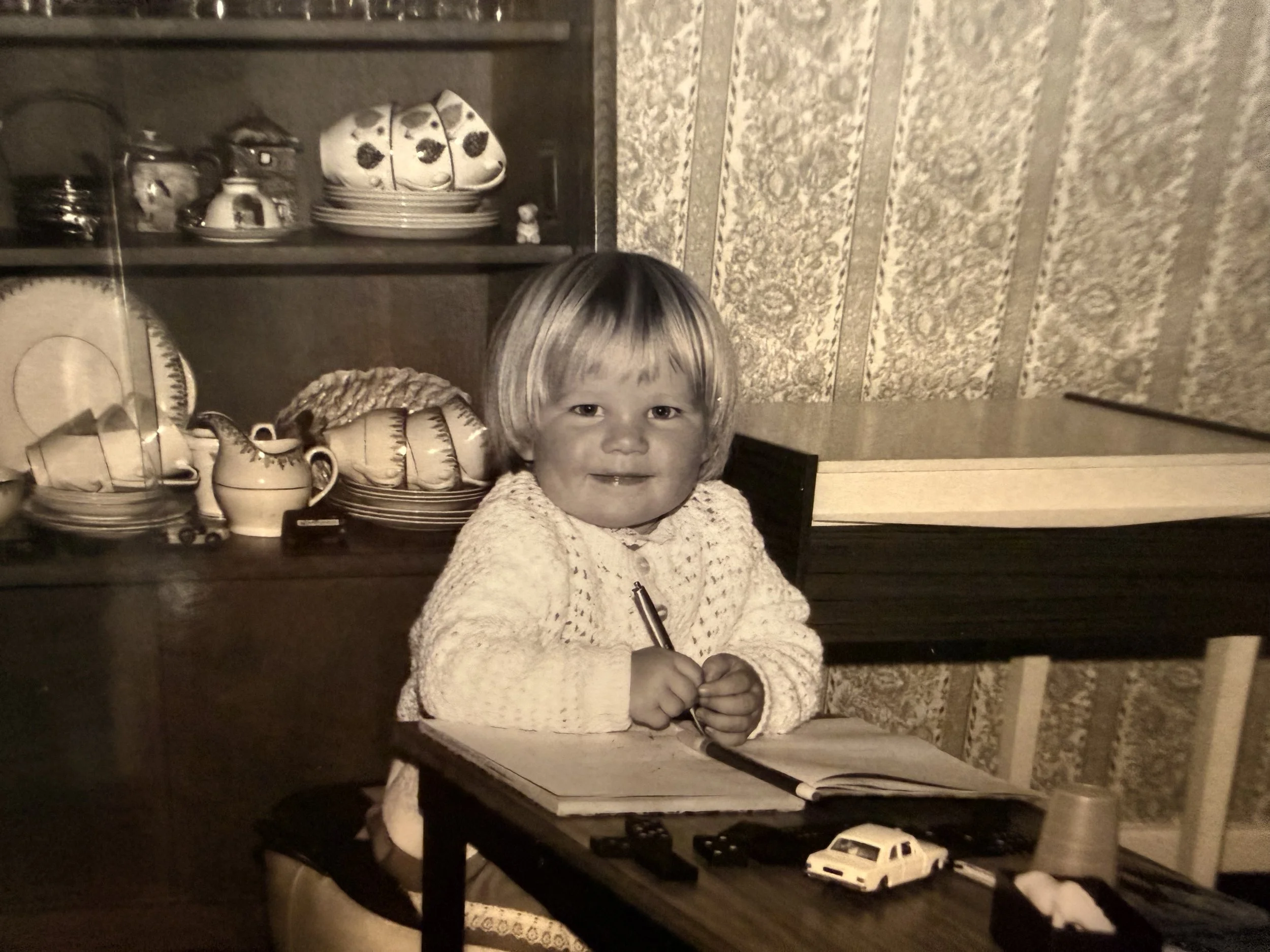 A young girl with short hair sitting at a table with a notebook, pen, toy car, and paper cups in front of her, smiling at the camera. A china cabinet with plates, teapots, and dishes is in the background.