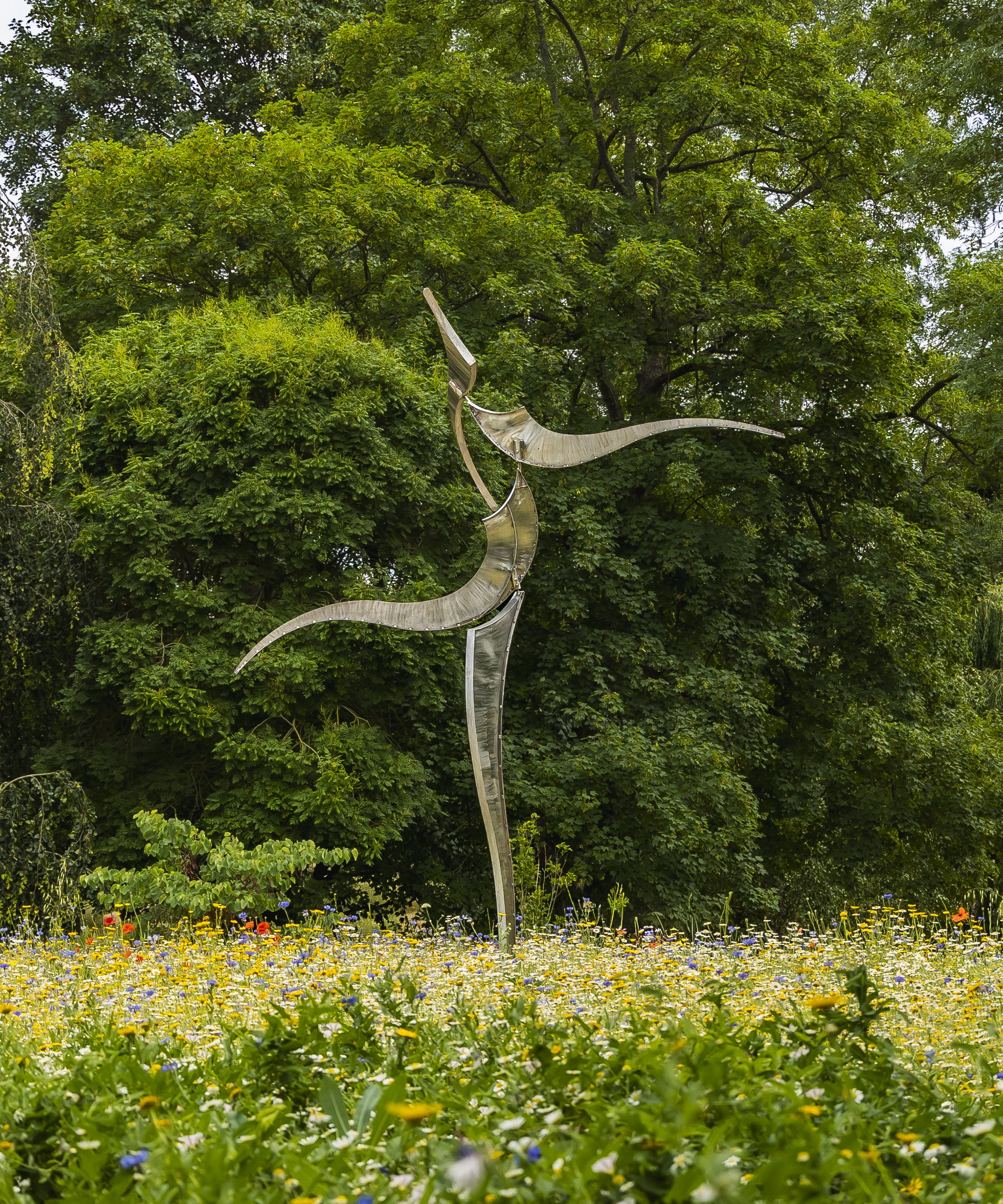 A tall metal sculpture resembling a winding garden path stands among a lush green tree and a vibrant flower field.