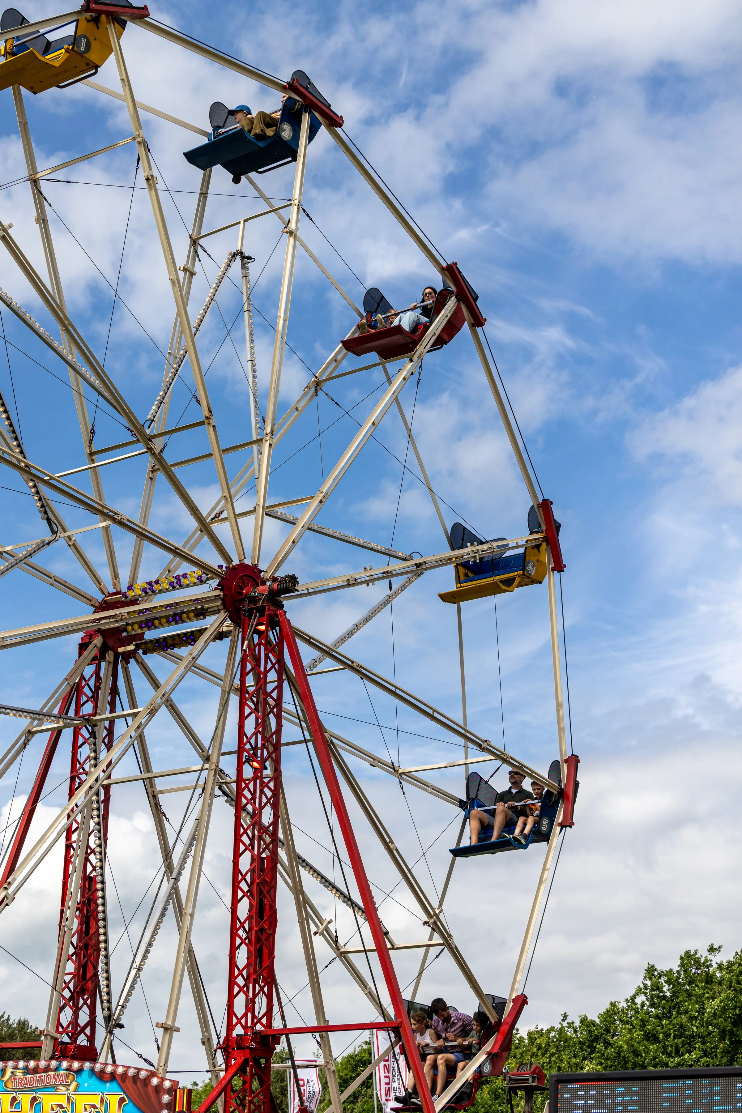 People riding a large Ferris wheel at an amusement park during daytime with a bright blue sky and some clouds.