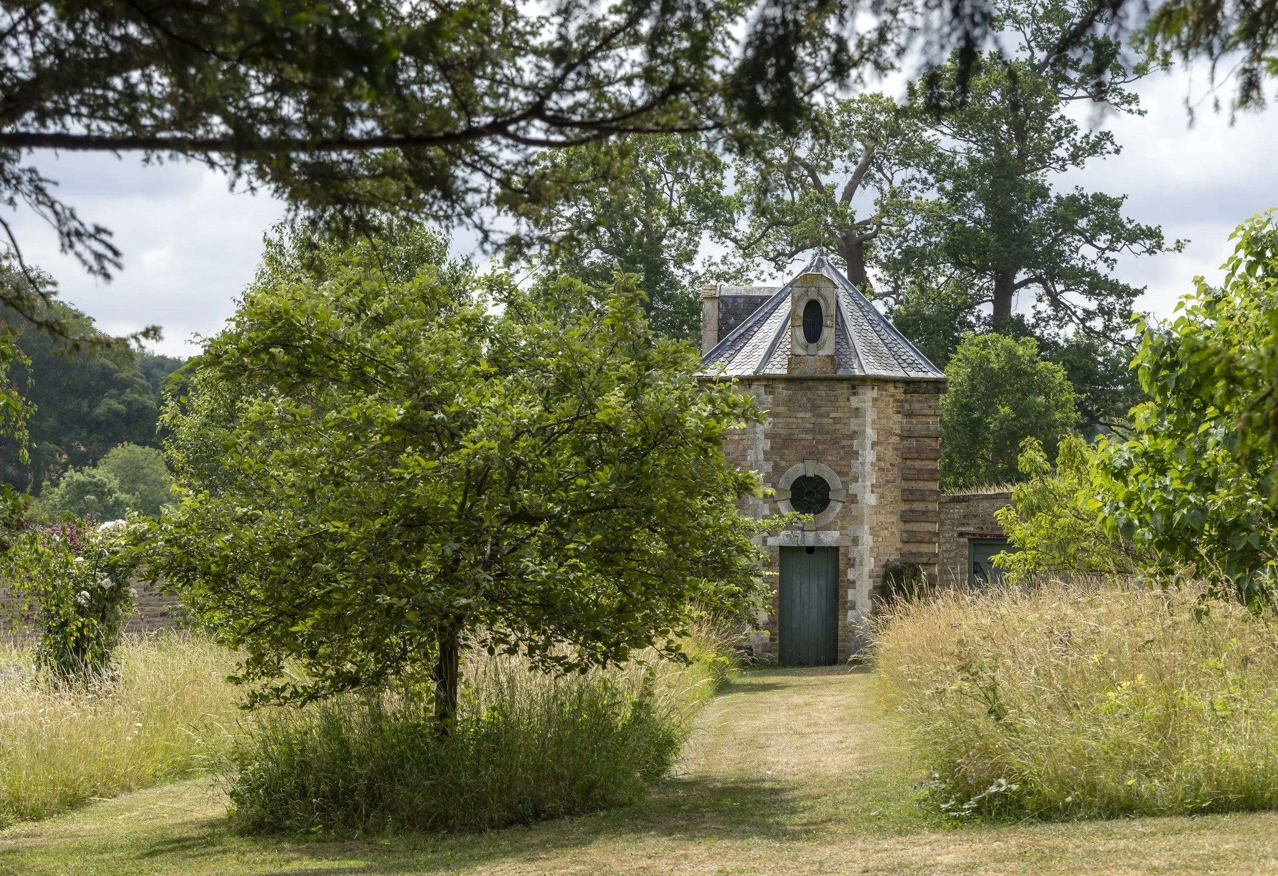 A small stone building with a conical roof surrounded by trees, shrubs, and a grassy pathway in a lush, green outdoor setting.
