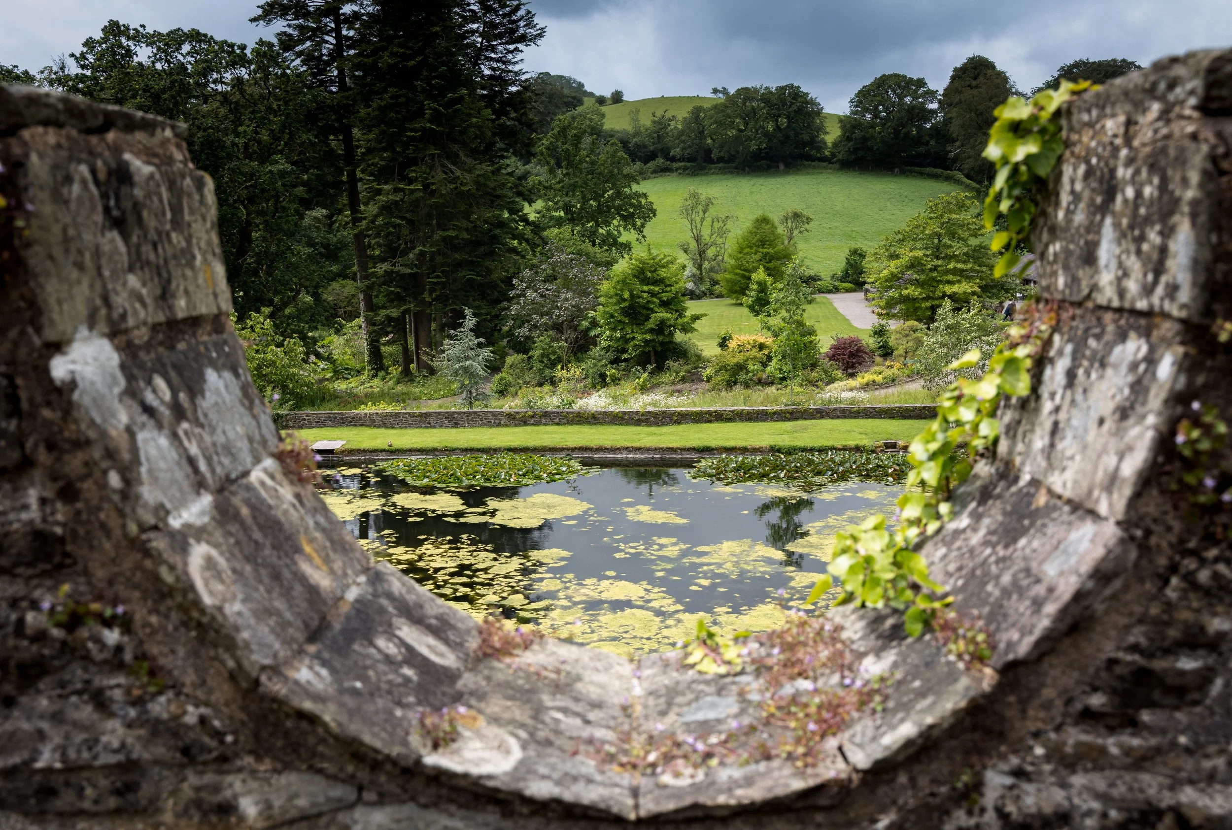 View of a lush green landscape with trees and a pond, framed by a stone opening.